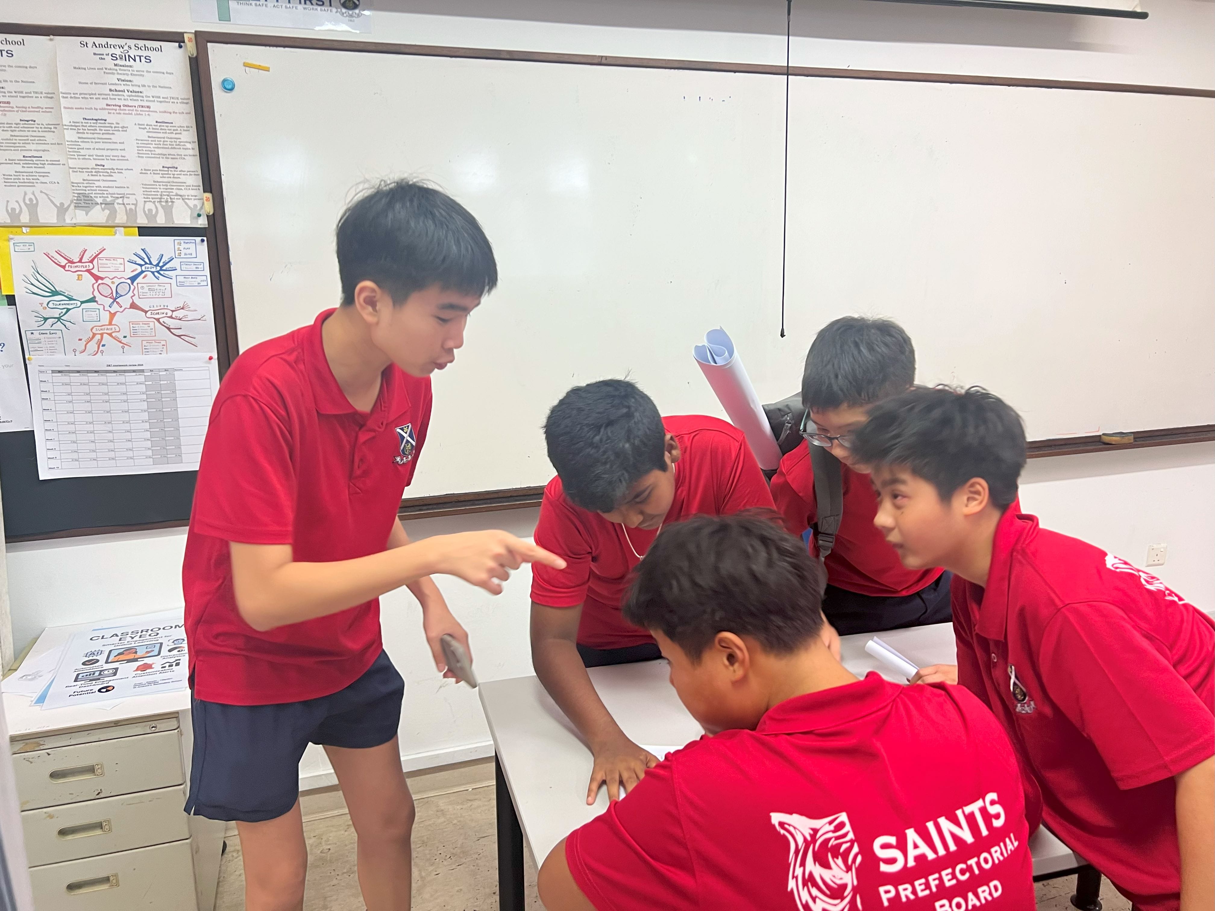 Five male students in red Saints school uniforms huddle around a desk in a classroom