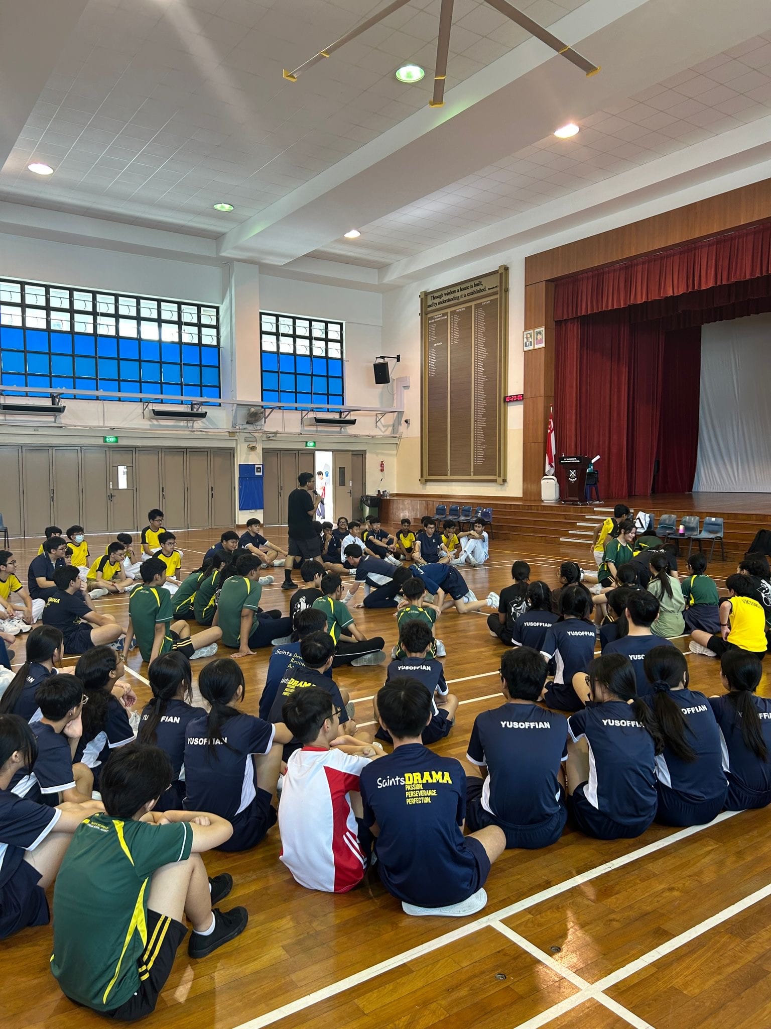 Students in school uniforms sit in a circle on a wooden hall floor, facing a standing instructor near a stage.