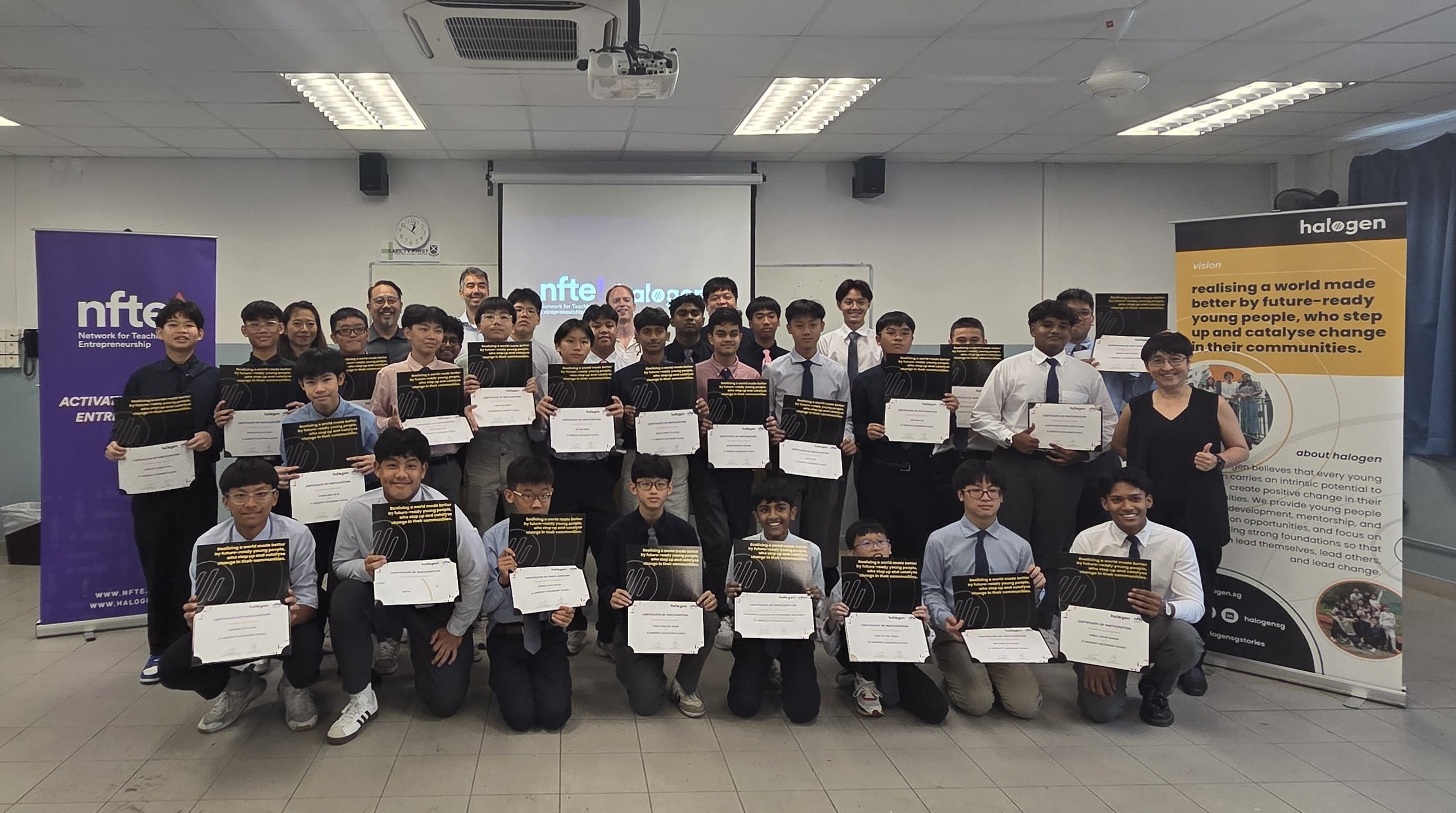 Large group of young male students holding certificates, posing indoors beside NFTE and Halogen banners.