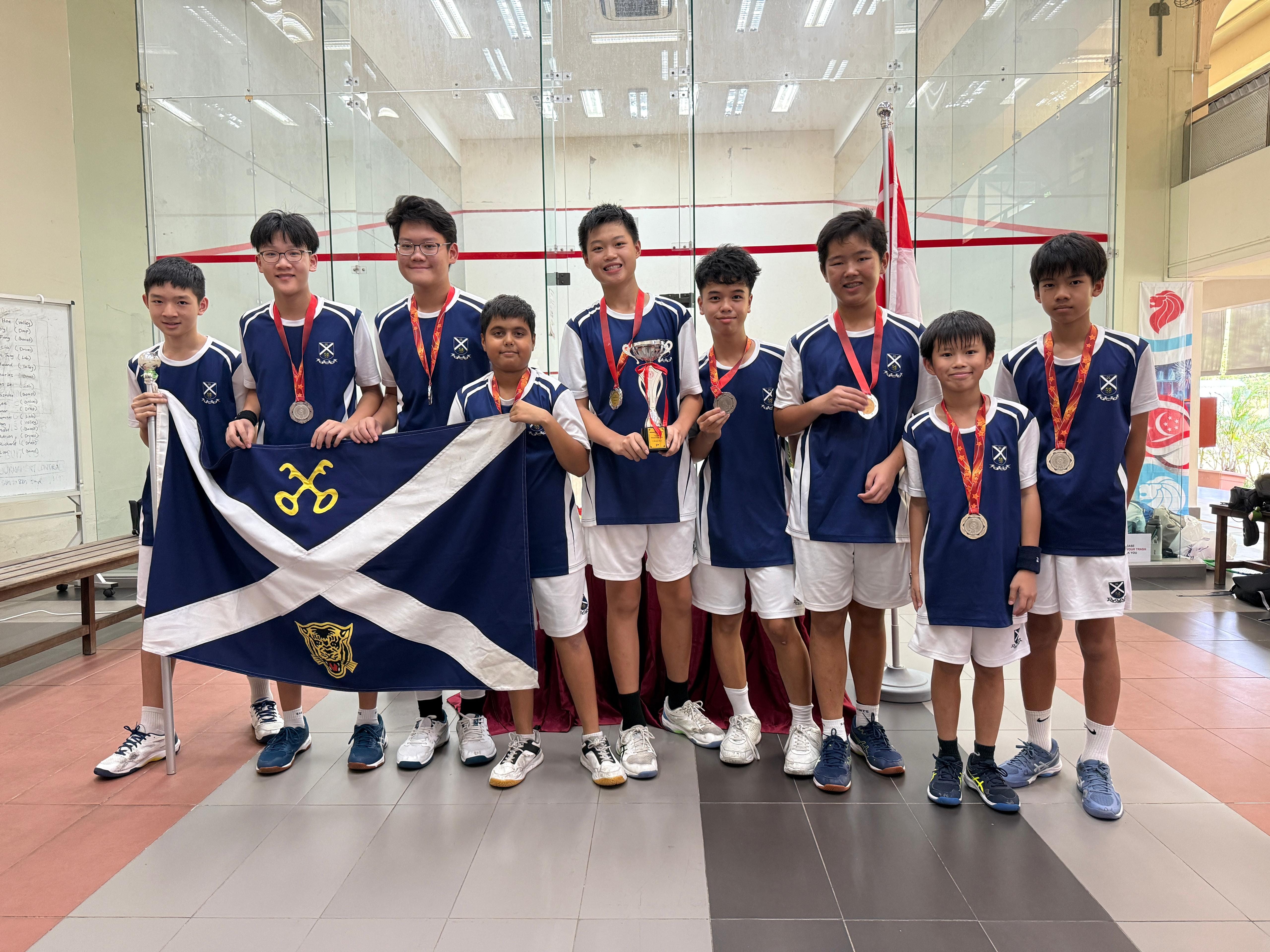 Nine boys in matching blue uniforms pose with medals, a trophy, and a navy flag inside a squash court.