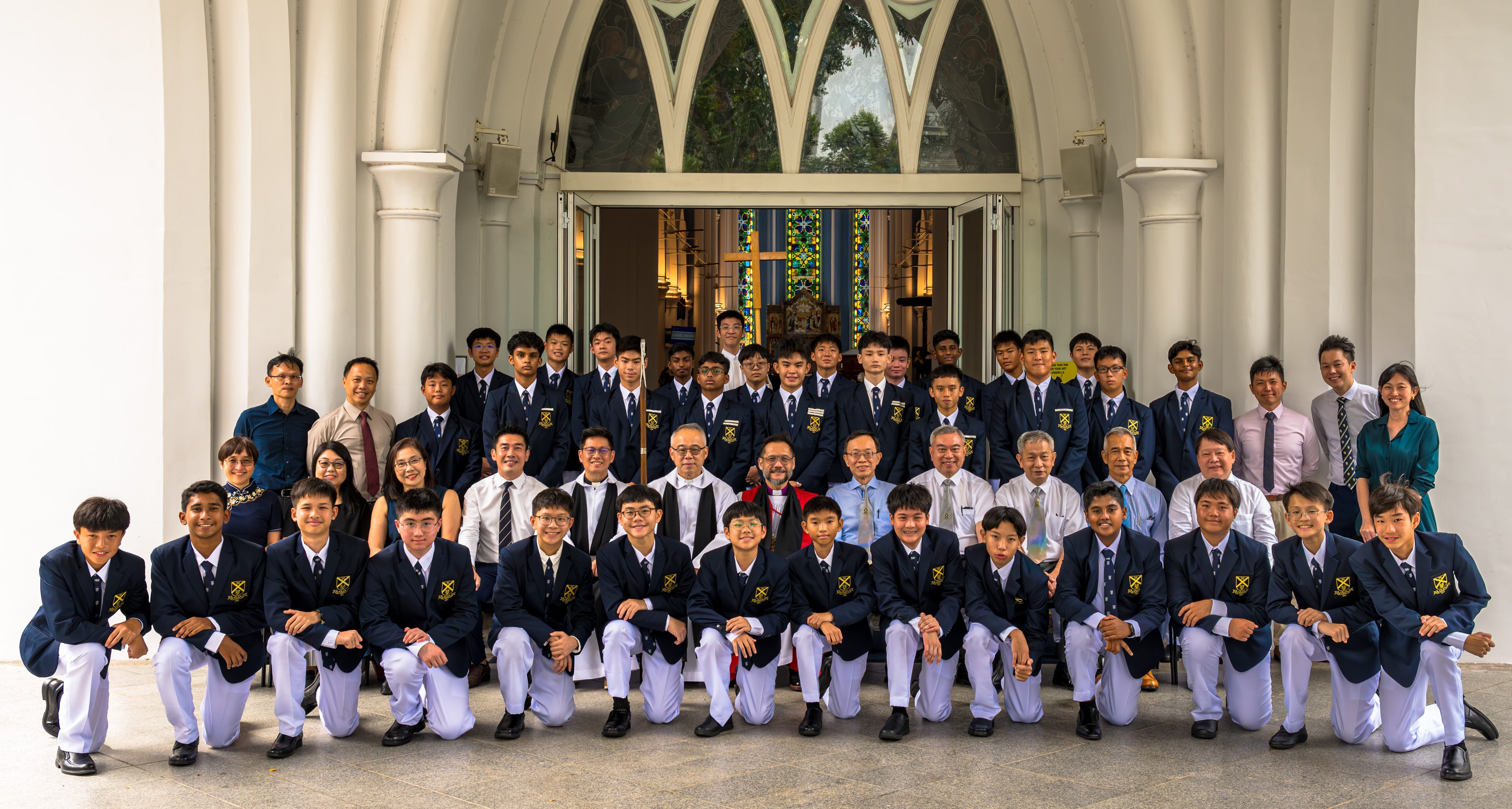 Large group of students in blazers and white trousers posing with adults outside a church entrance.