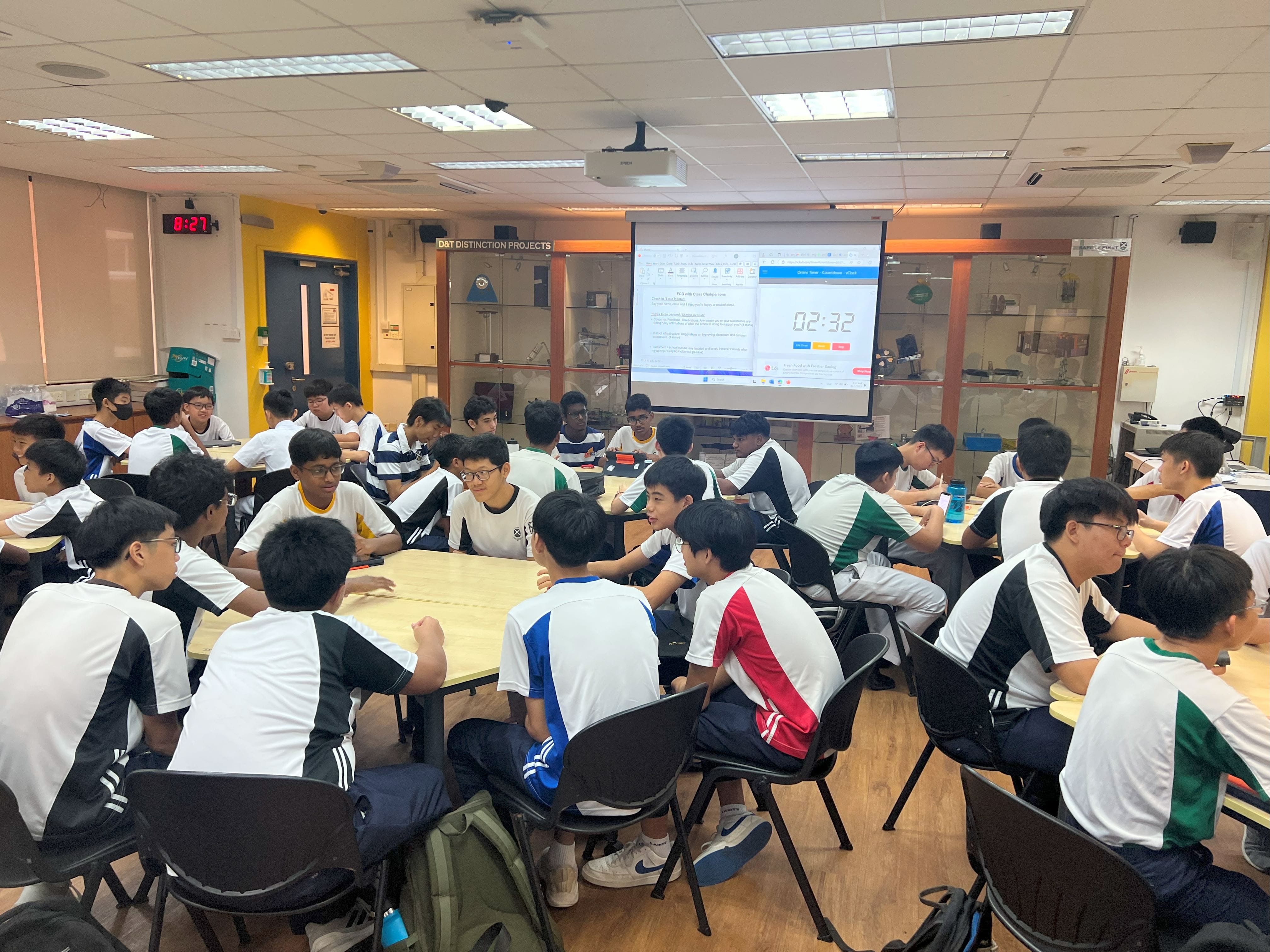 Students sit at grouped tables in a classroom facing a projected screen showing a countdown timer.