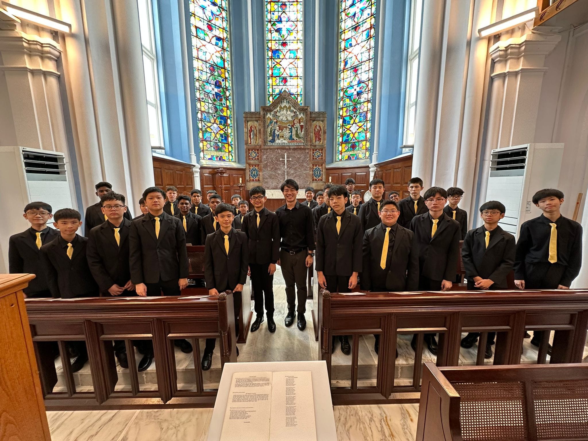 A group of young males in black suits with yellow ties stand in a church with stained glass windows behind them.