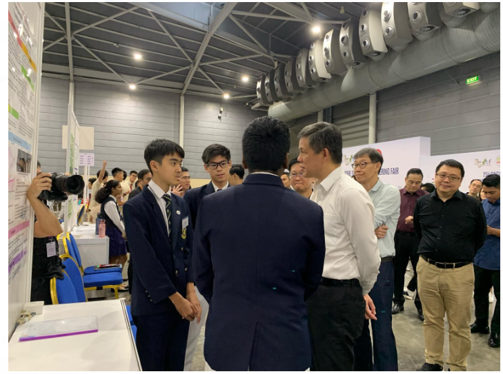 Students in school uniforms present their project to adults at an indoor science or engineering fair.
