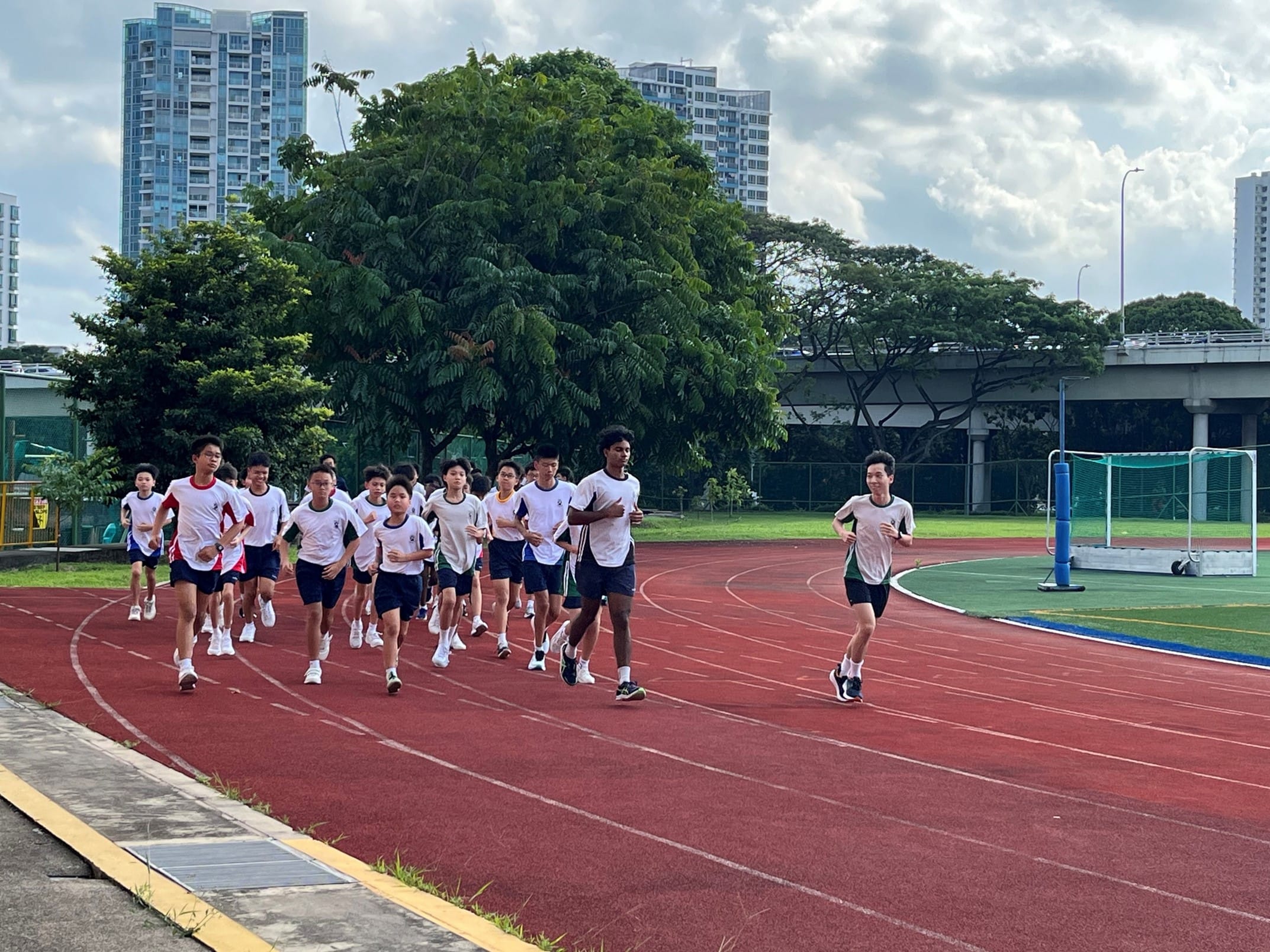 A group of students in white shirts and dark shorts running on a red athletic track with trees and buildings behind.