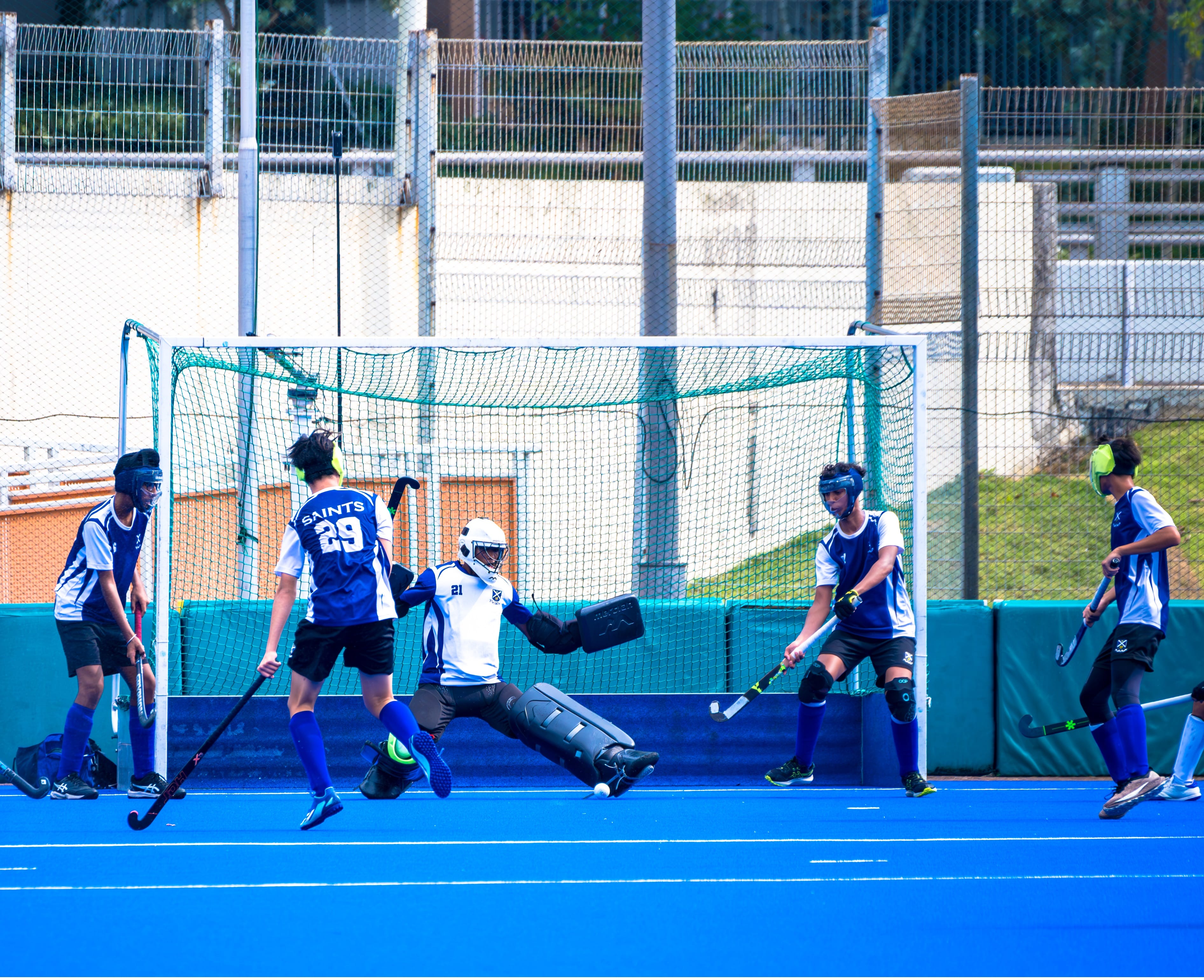Young players compete in a field hockey match, with a goalkeeper diving to block a shot at a green-netted goal.