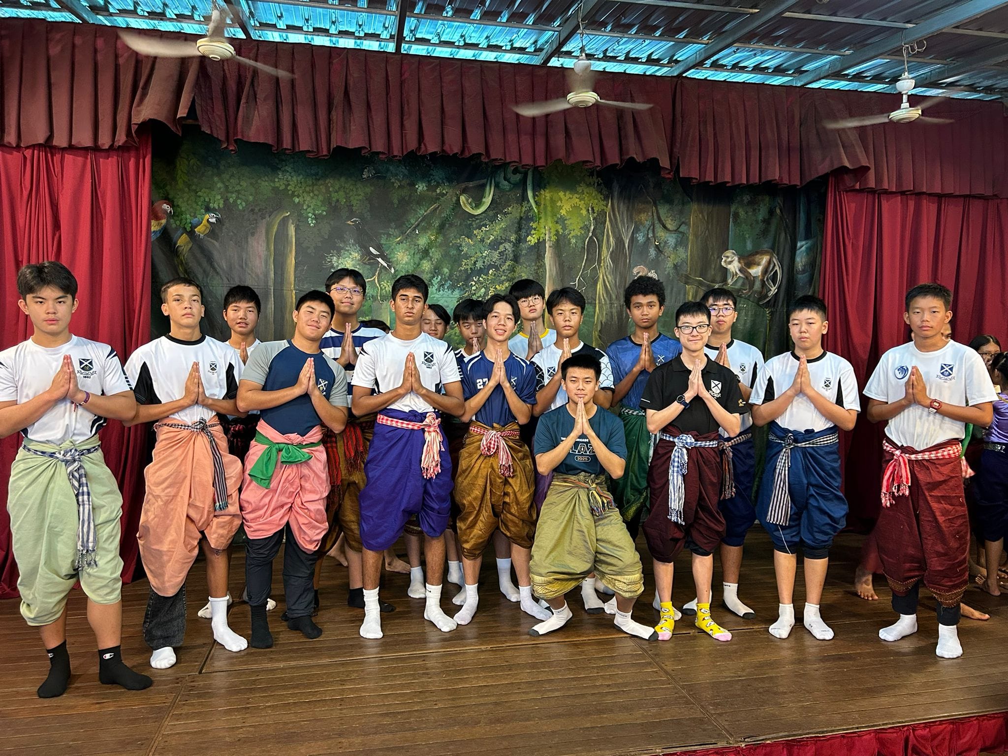 A group of teenage boys in traditional pants press their palms together on a wooden stage.