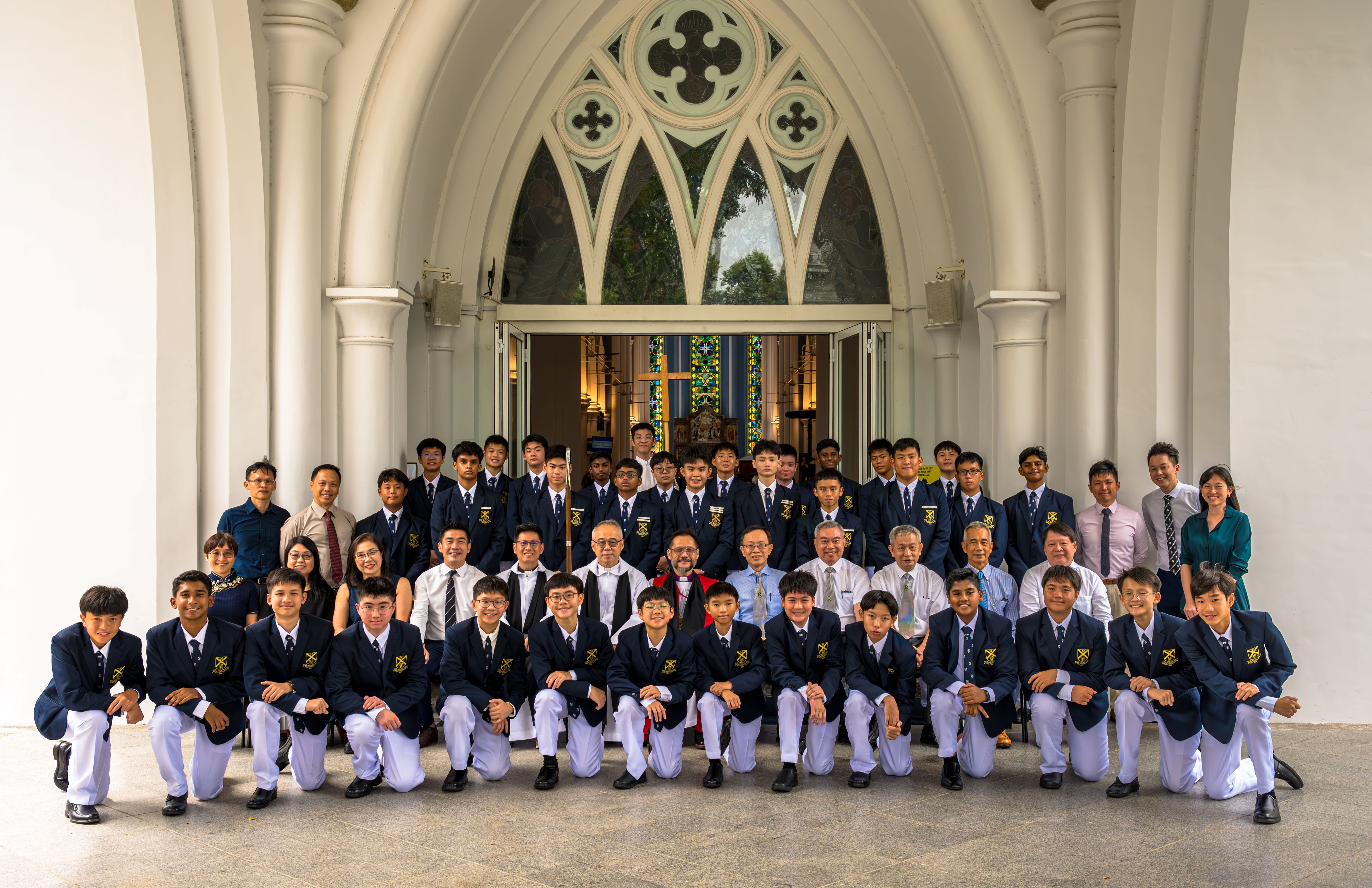 A large group of students in navy blazers and white trousers pose with adults at a church entrance.