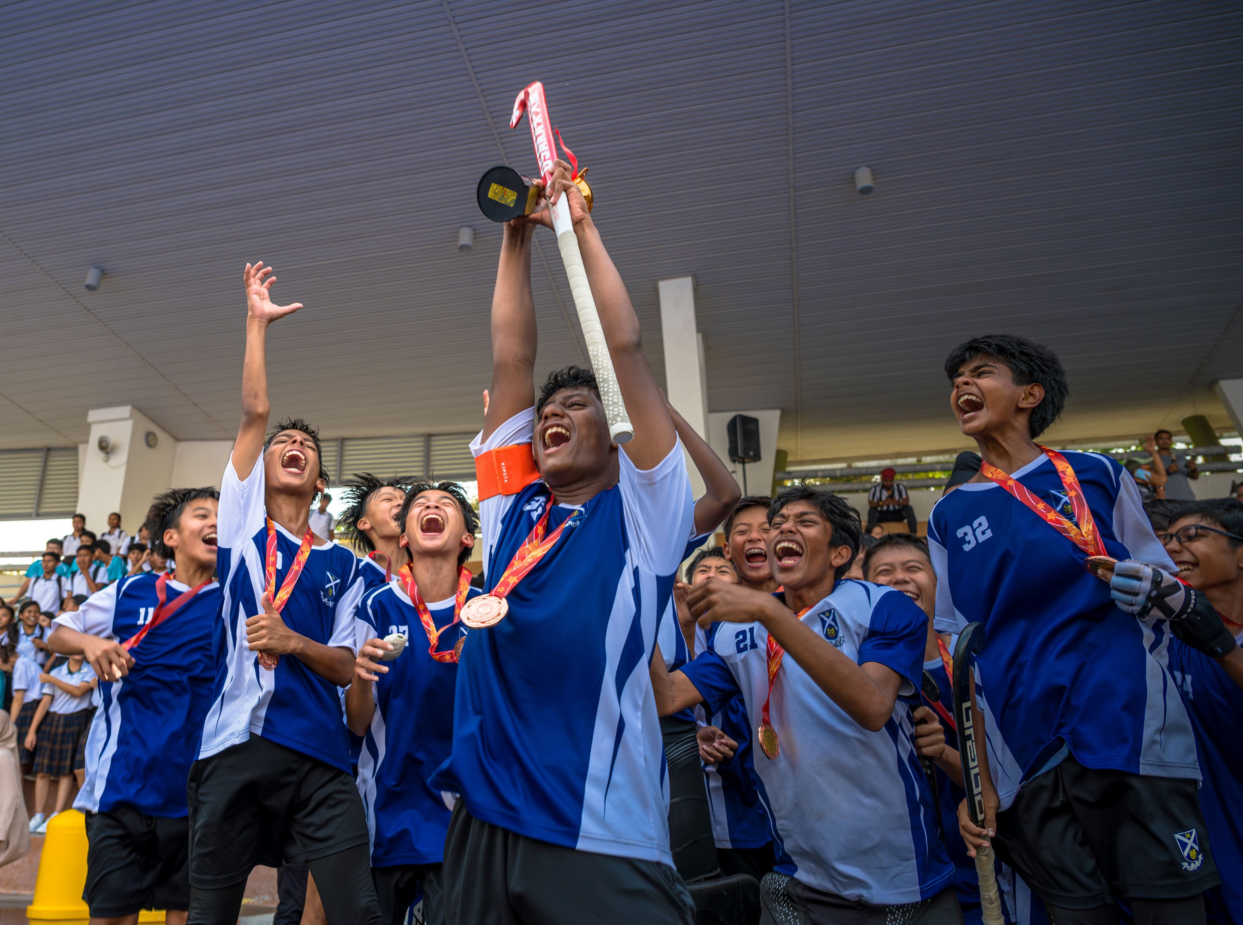 Young hockey players in blue jerseys celebrate joyfully as one raises a trophy overhead, with medals around their necks.