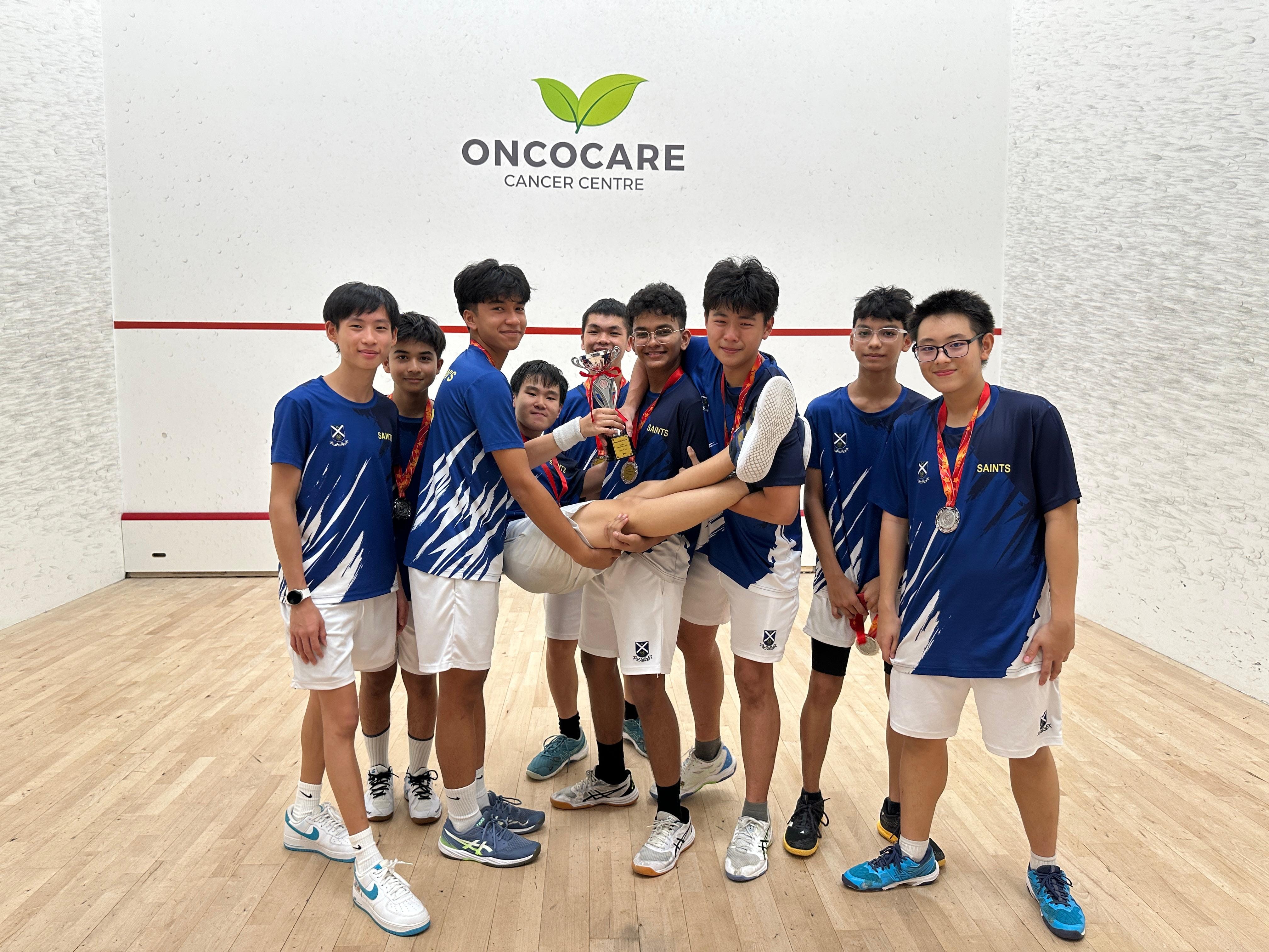 A group of boys in blue Saints jerseys with medals pose on a squash court holding a trophy.