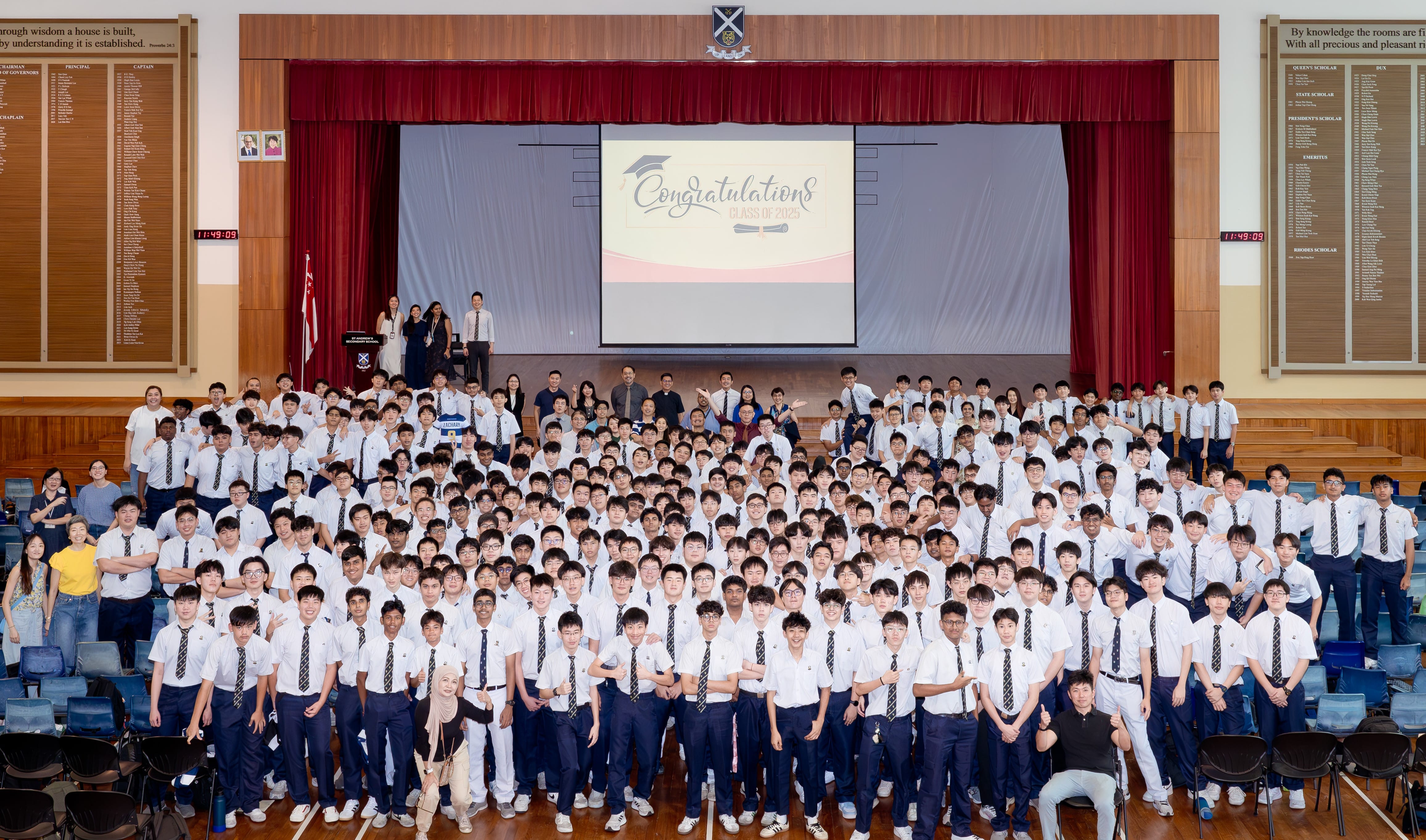 Large group of students in white shirts and ties posing in a school hall with a Congratulations Class of 2025 screen.