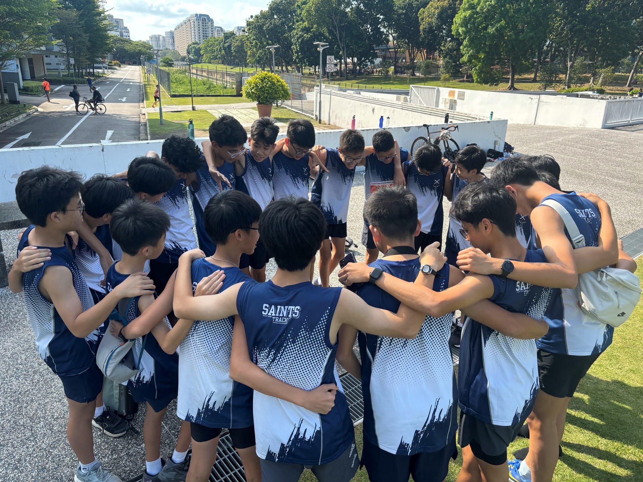 A group of young male athletes in Saints Track uniforms huddle together outdoors near a road and park.