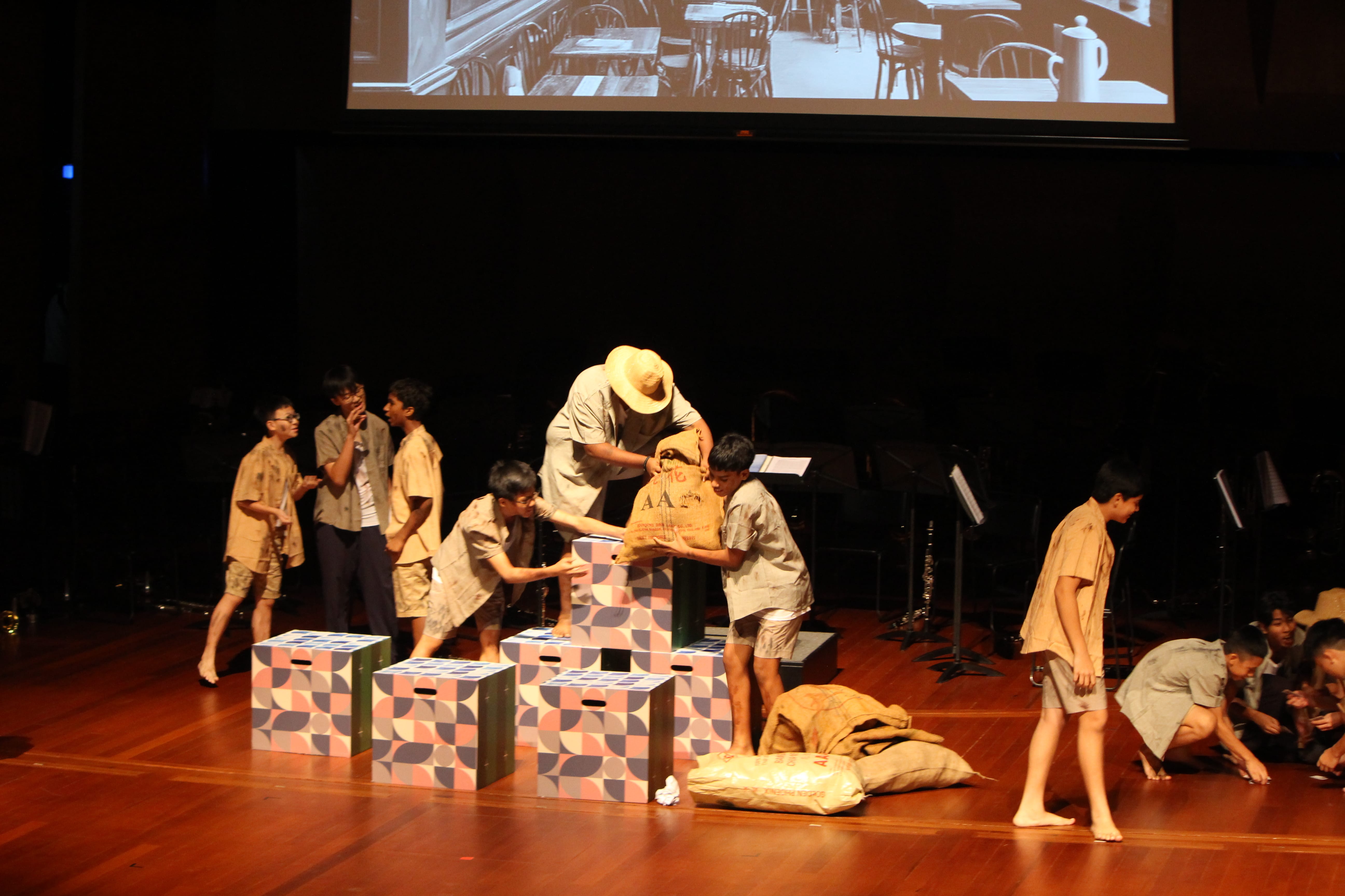 Some student performers moving large sacks on a stage with some square cardboard boxes