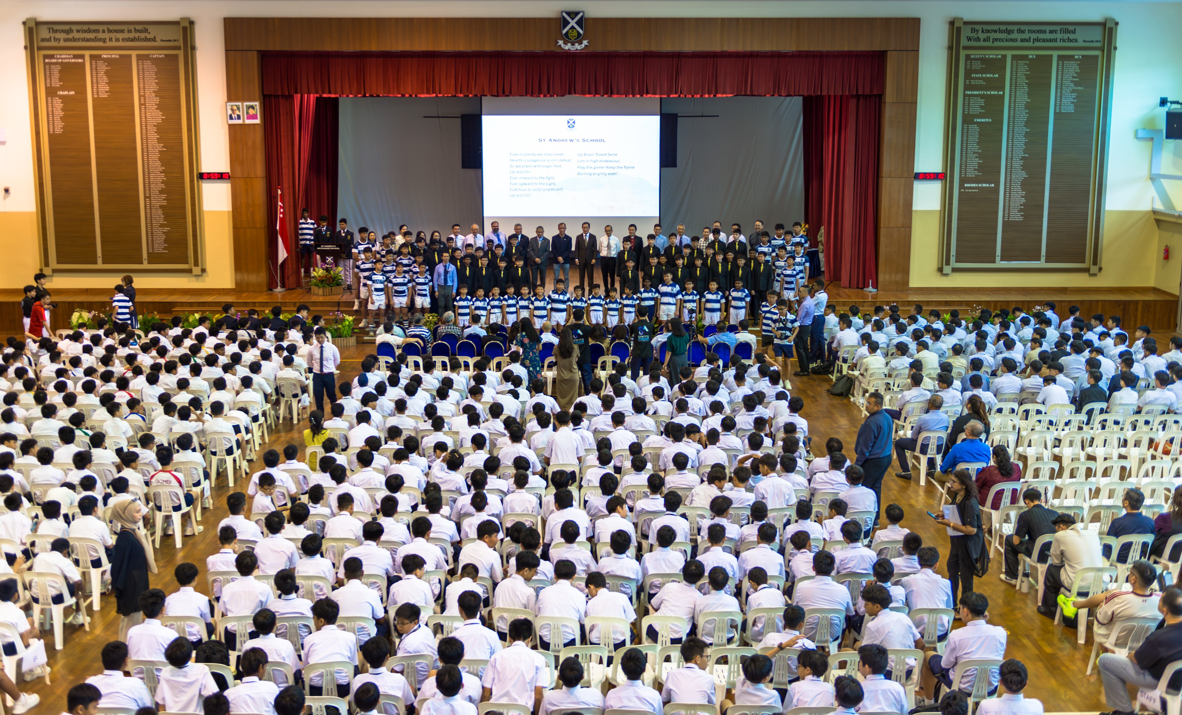 Large school assembly hall filled with students in white uniforms seated in rows, facing a stage with a projected screen.