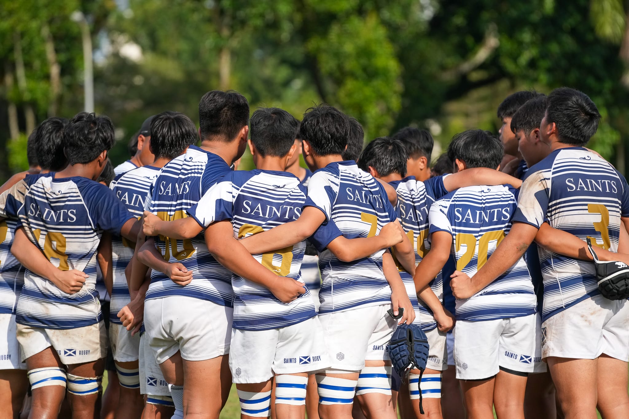 A rugby team wearing blue and white Saints jerseys huddle together with arms around each other outdoors.