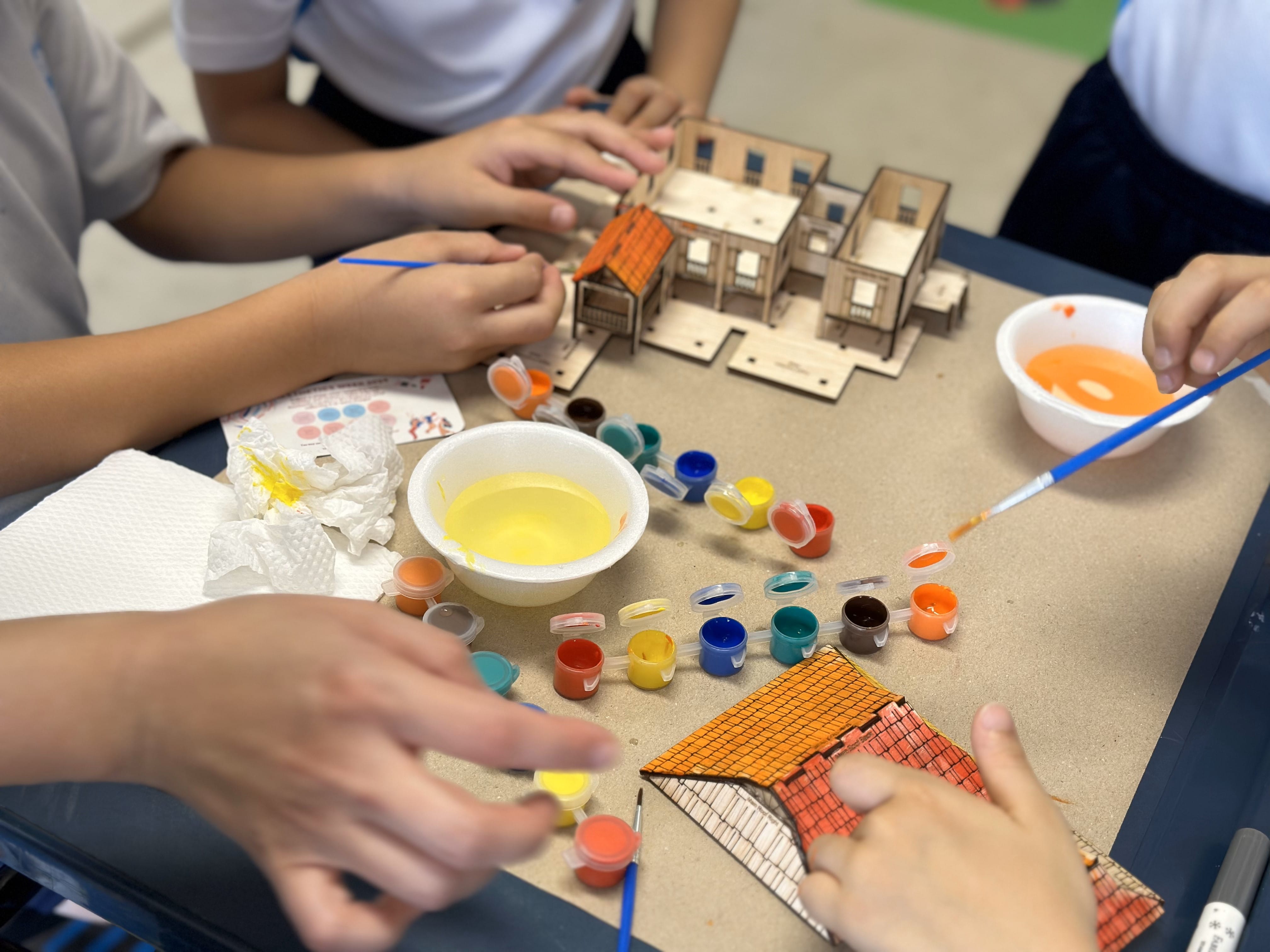 Children painting a small wooden model house using colorful paint pots and brushes on a table.