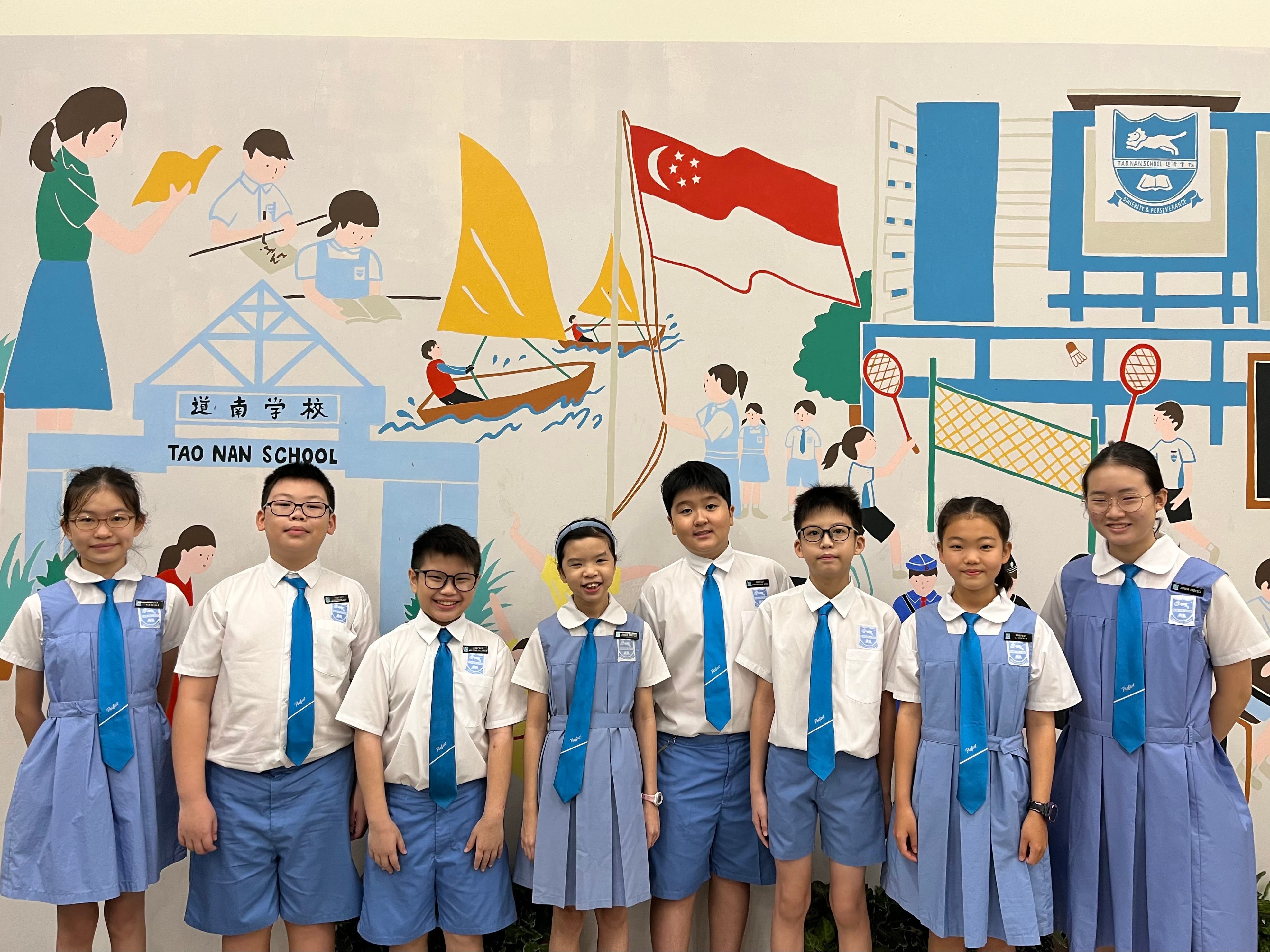 Eight students in Tao Nan School uniforms stand smiling in front of a colorful school mural.