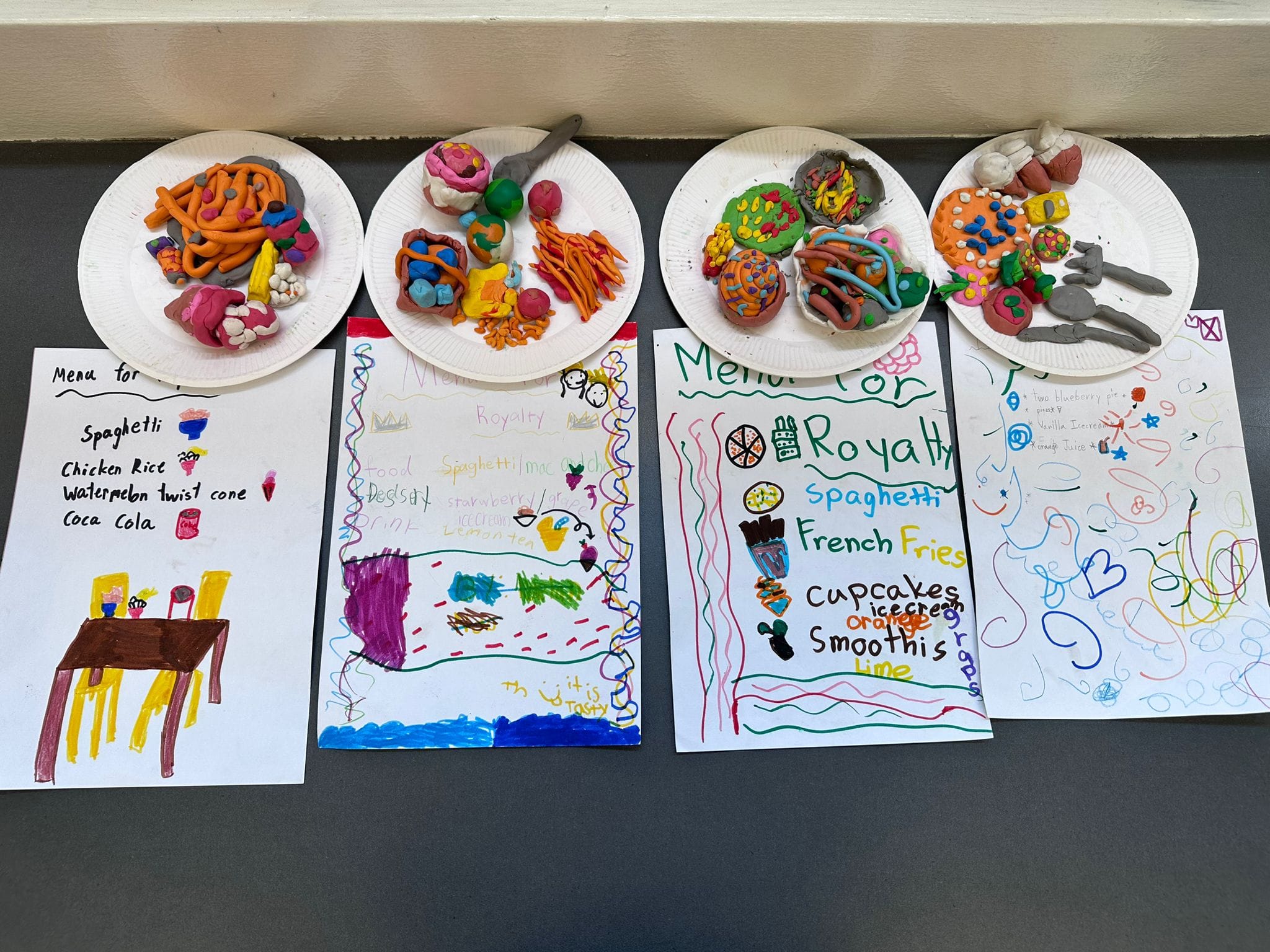 Four children's handwritten menus with colorful clay food models on paper plates displayed on a gray surface.