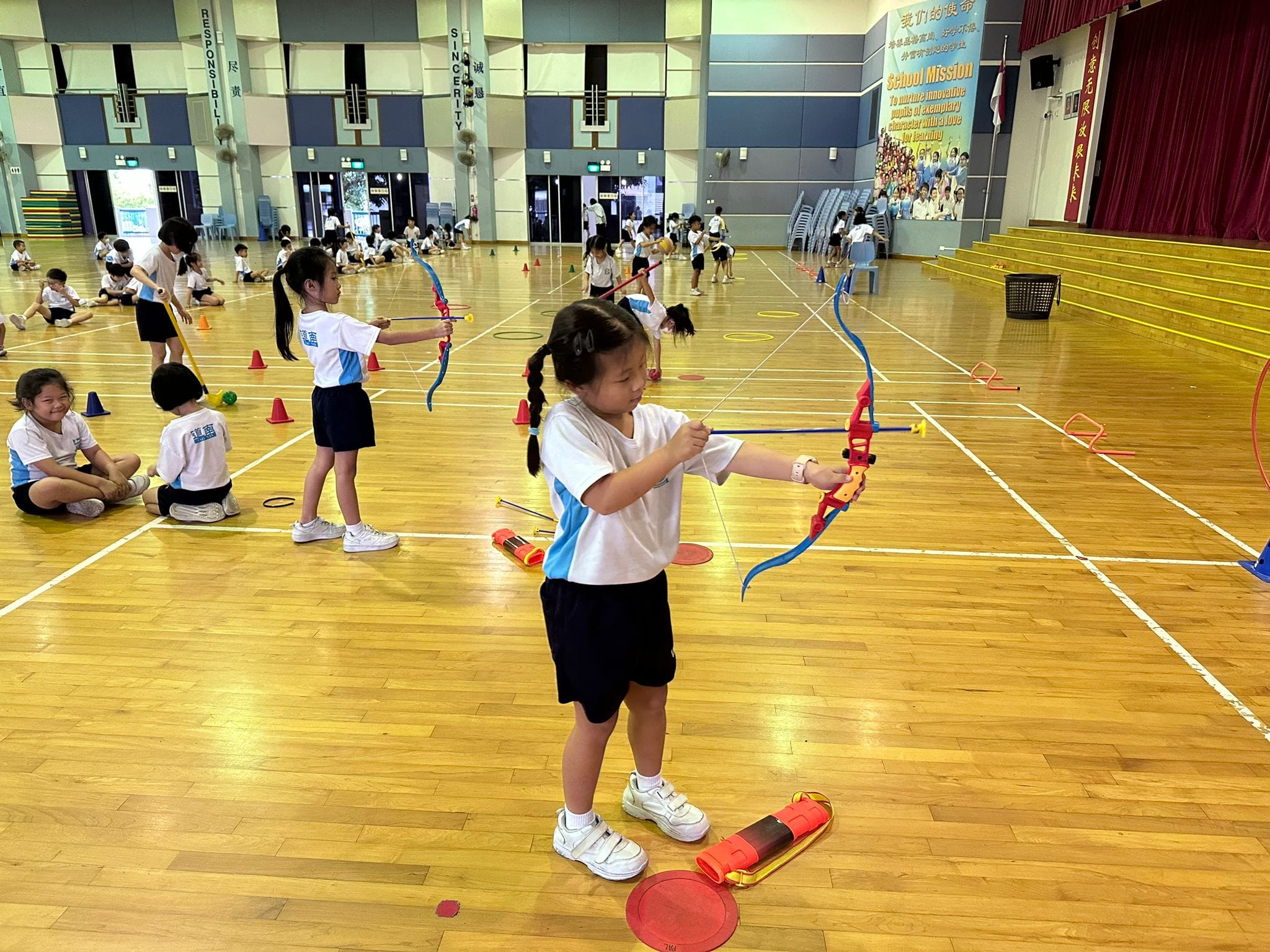 Young students in school uniforms practice archery with toy bows inside a gymnasium with cones and hoops on the floor.