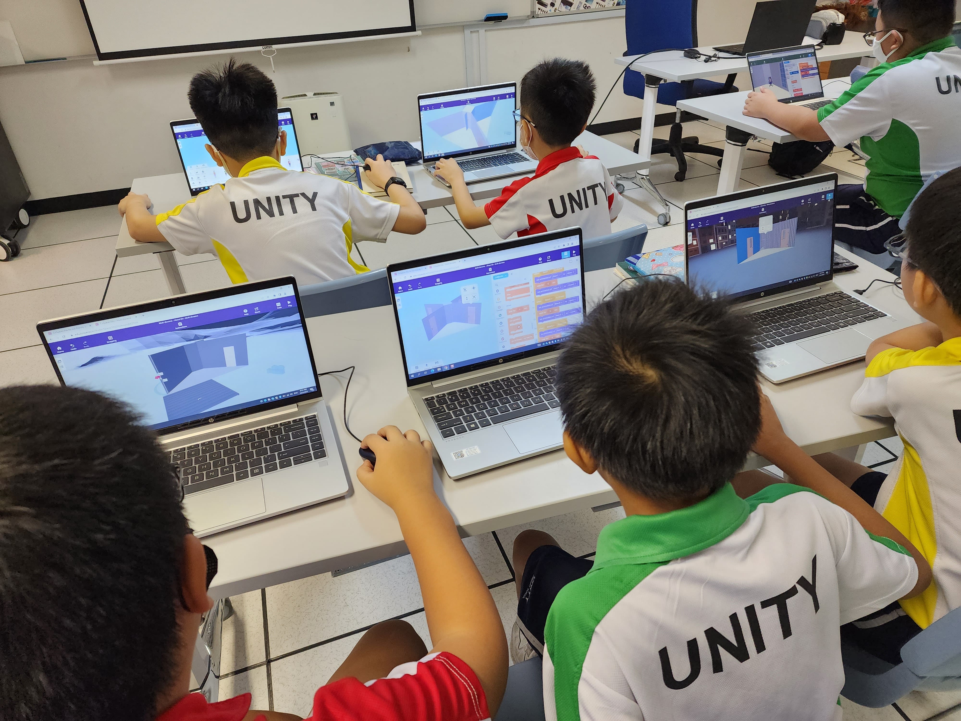 Several children wearing Unity shirts work on laptops displaying 3D design and coding software in a classroom.