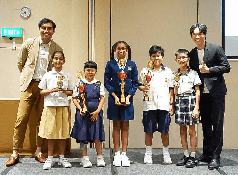 Four children in school uniforms hold trophies while posing with two adult men in a hall.