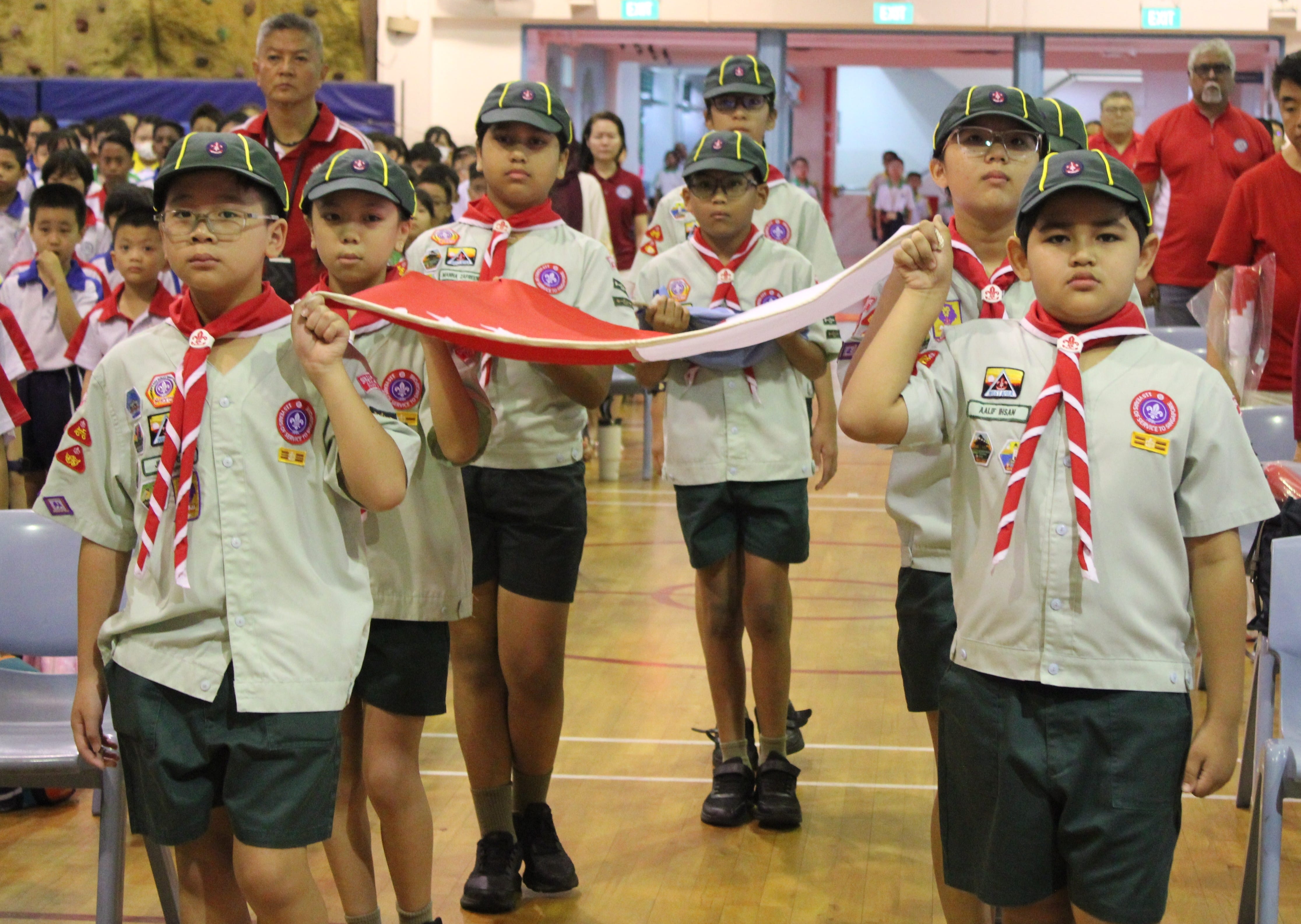 Young scouts in uniform with red neckerchiefs carry a folded red and white flag during an indoor ceremony.