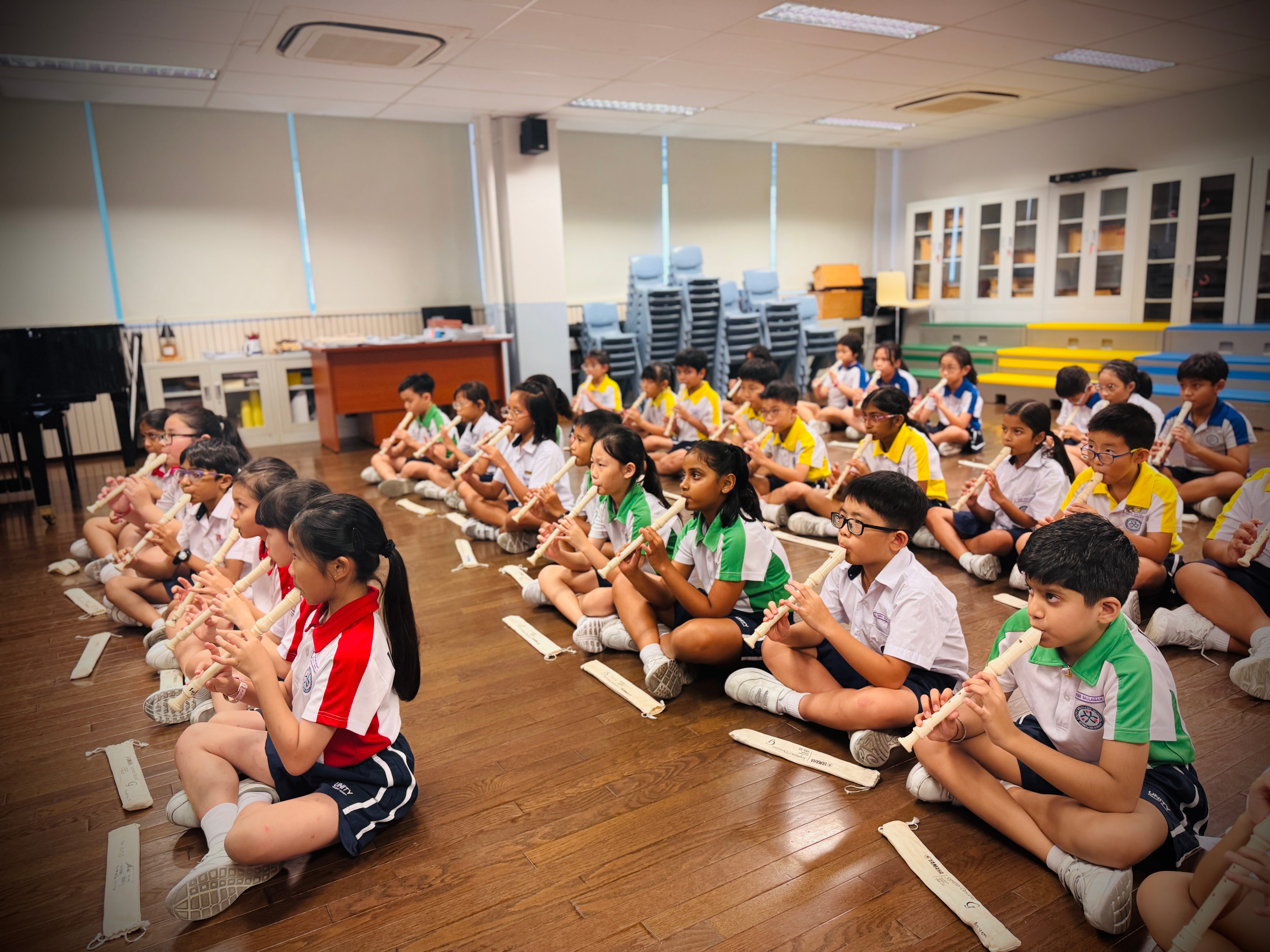 Students sit cross-legged on a classroom floor playing recorders, with stacked chairs and cabinets behind.