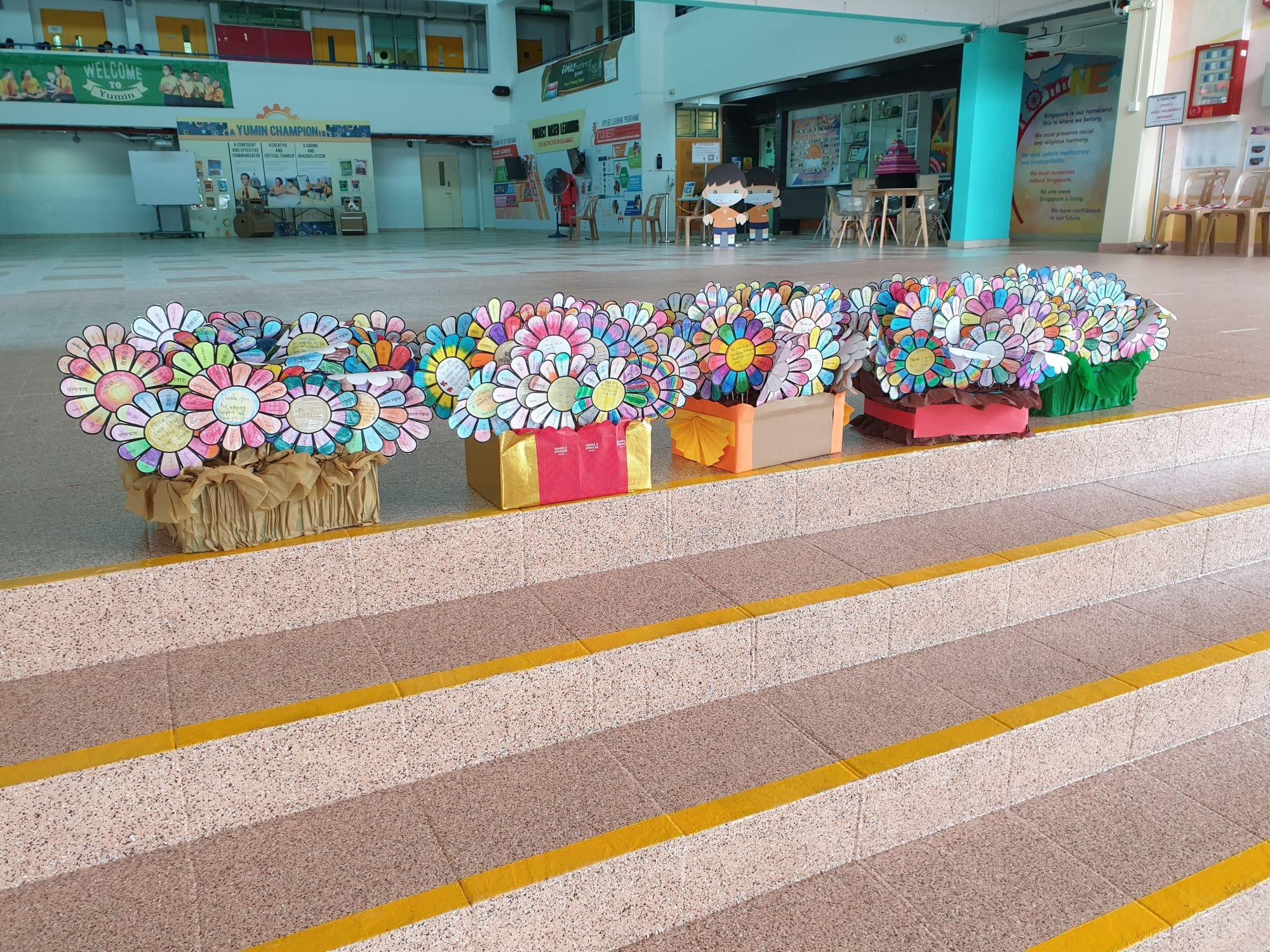 Colorful handmade paper flowers with written petals displayed in decorated boxes in a school hall.