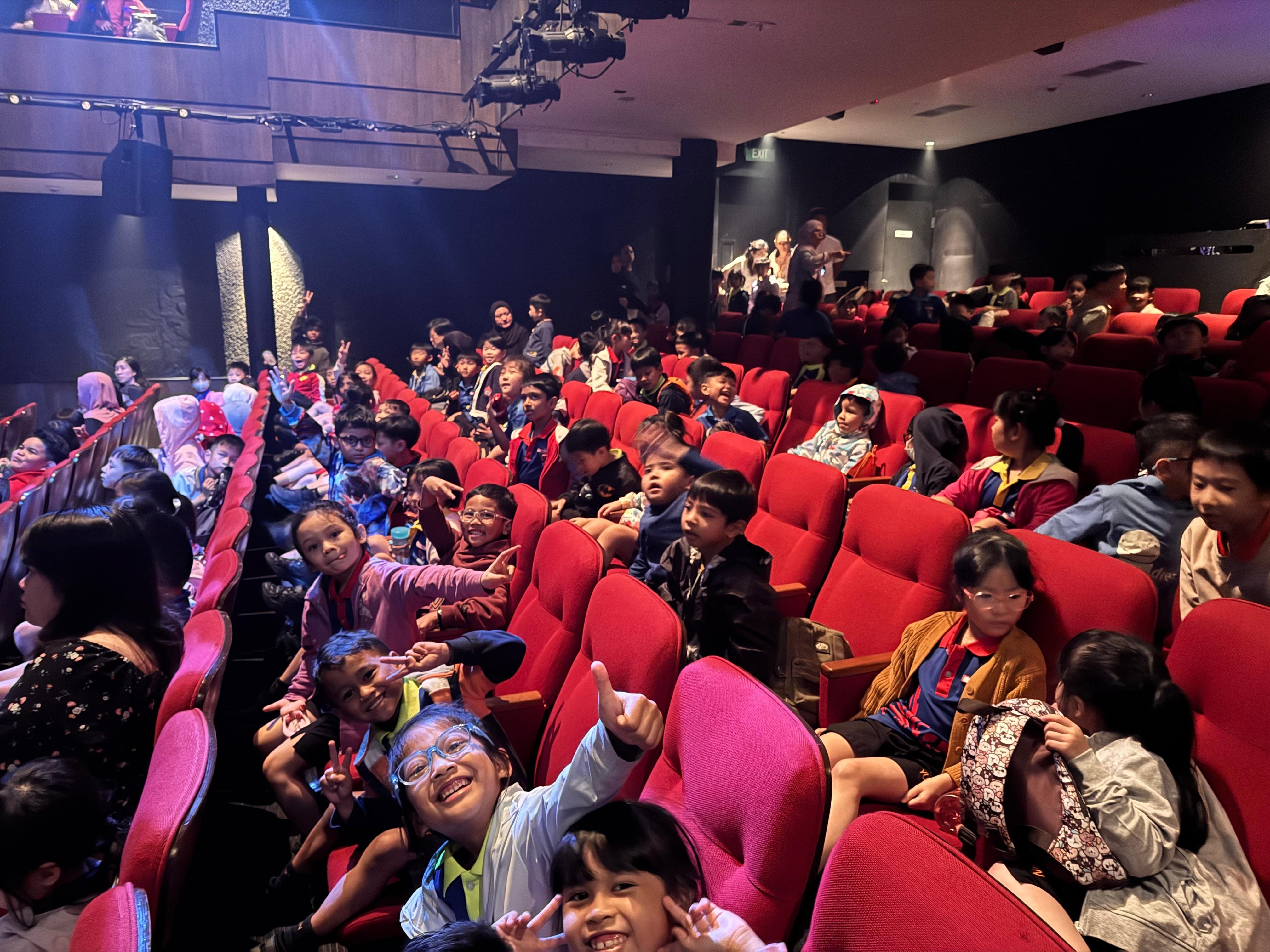 A group of children seated in red theater seats, smiling towards the camera.