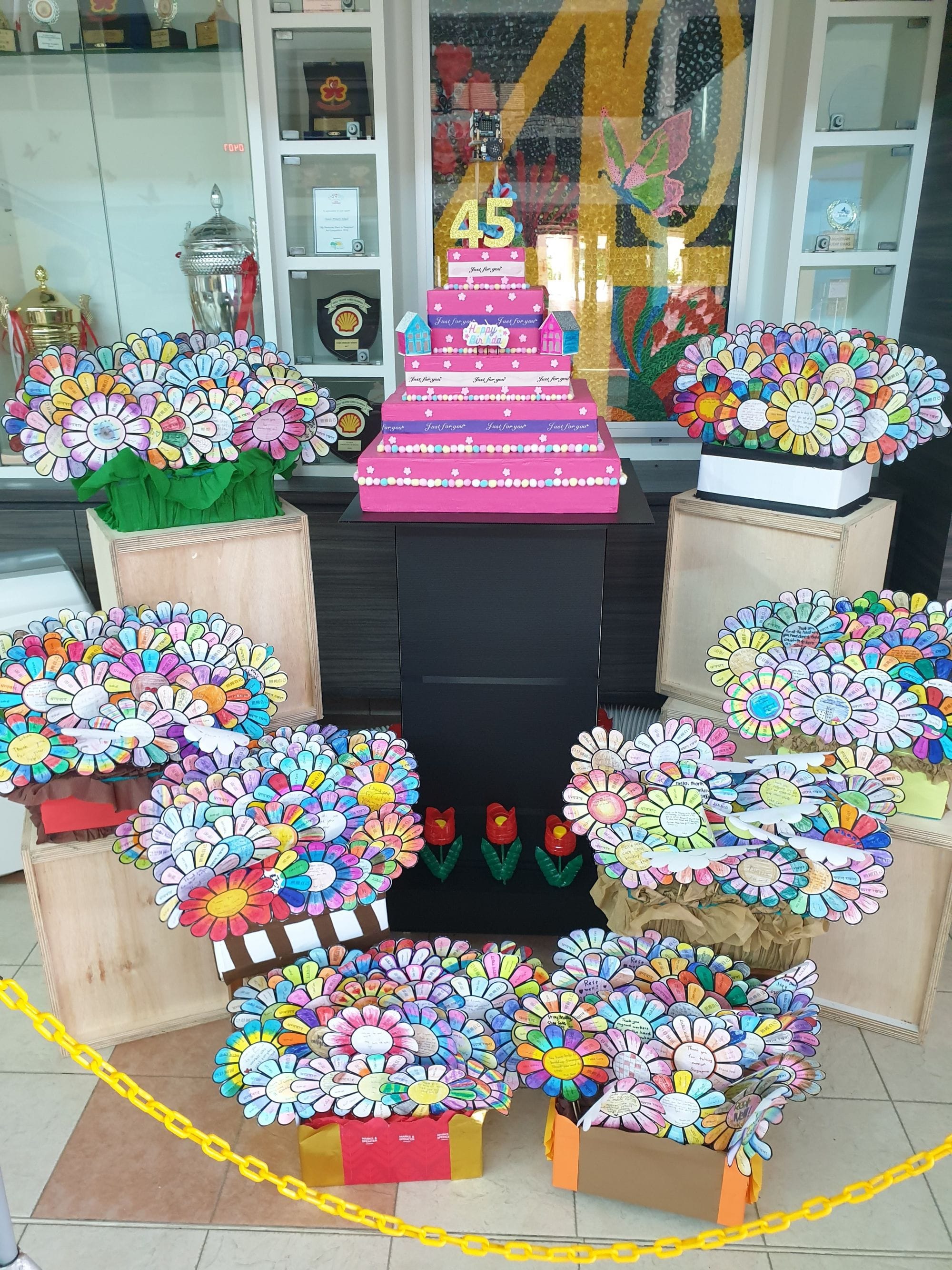 A colorful display of handmade paper flowers surrounds a pink tiered birthday cake decorated with the number 45.