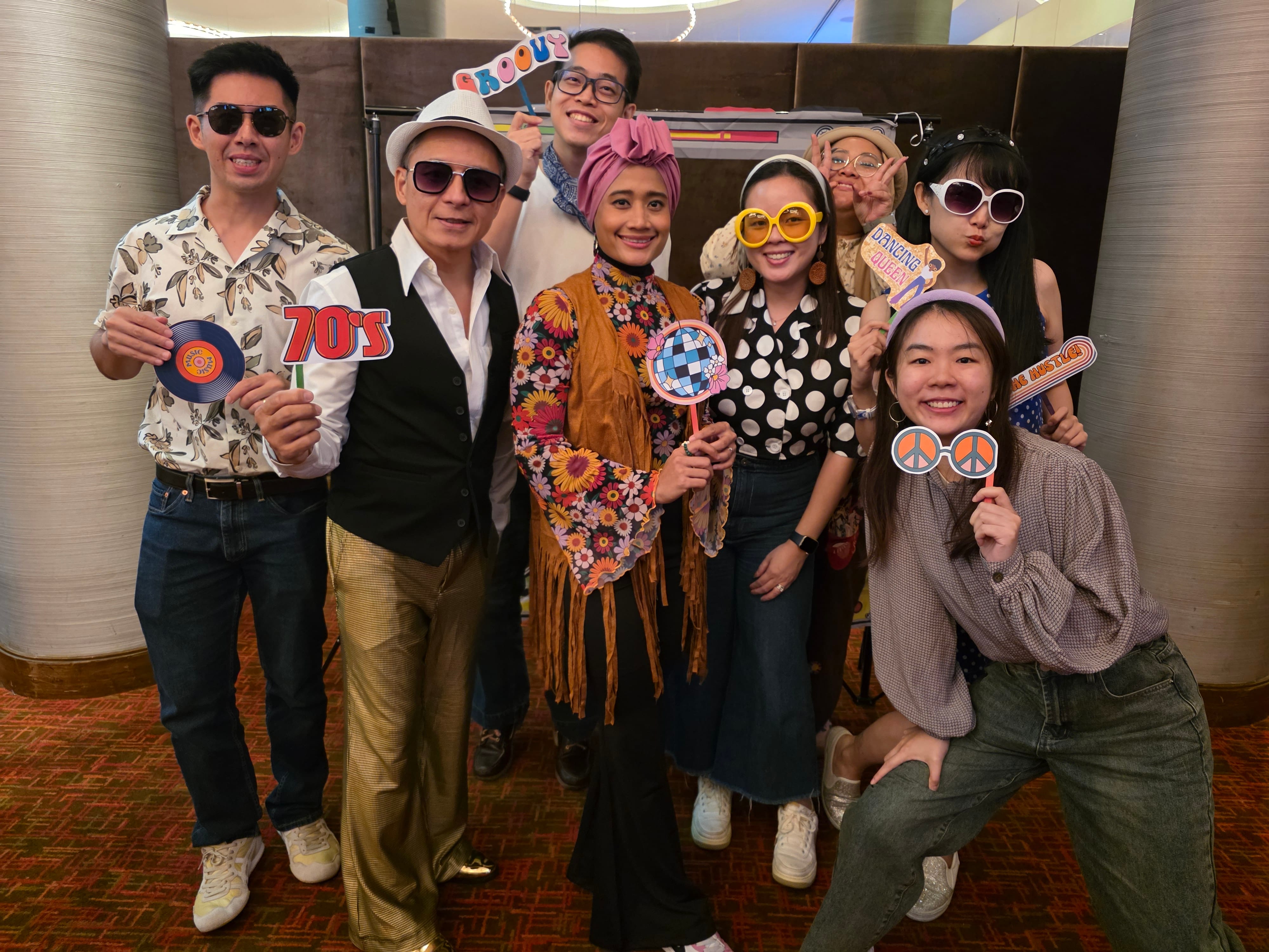 Eight people posing in 70s-themed costumes holding photo booth props like a vinyl record, disco ball, and peace sign glasses.