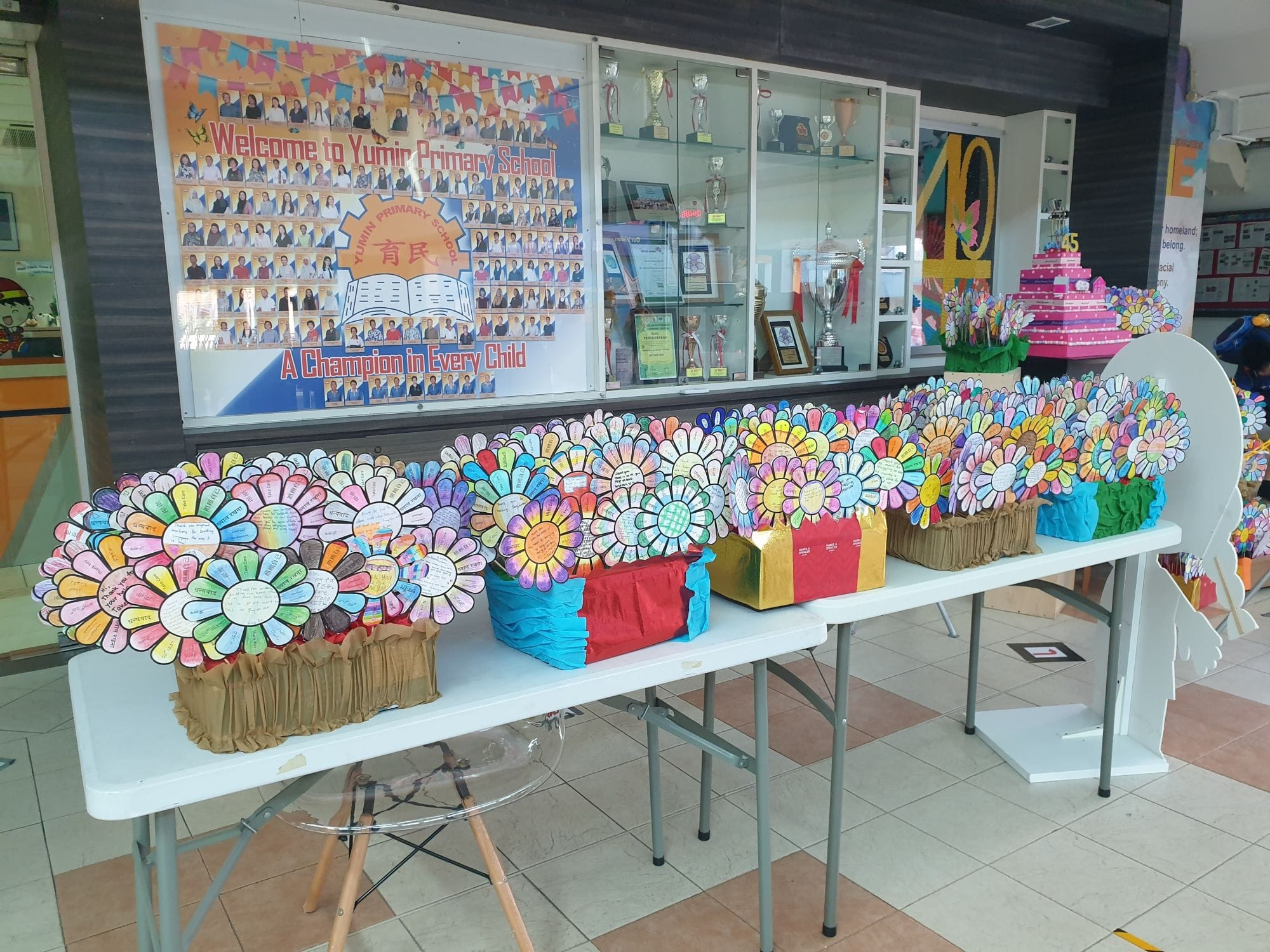 Colorful paper flower crafts with written messages displayed on tables in Yumin Primary School lobby.