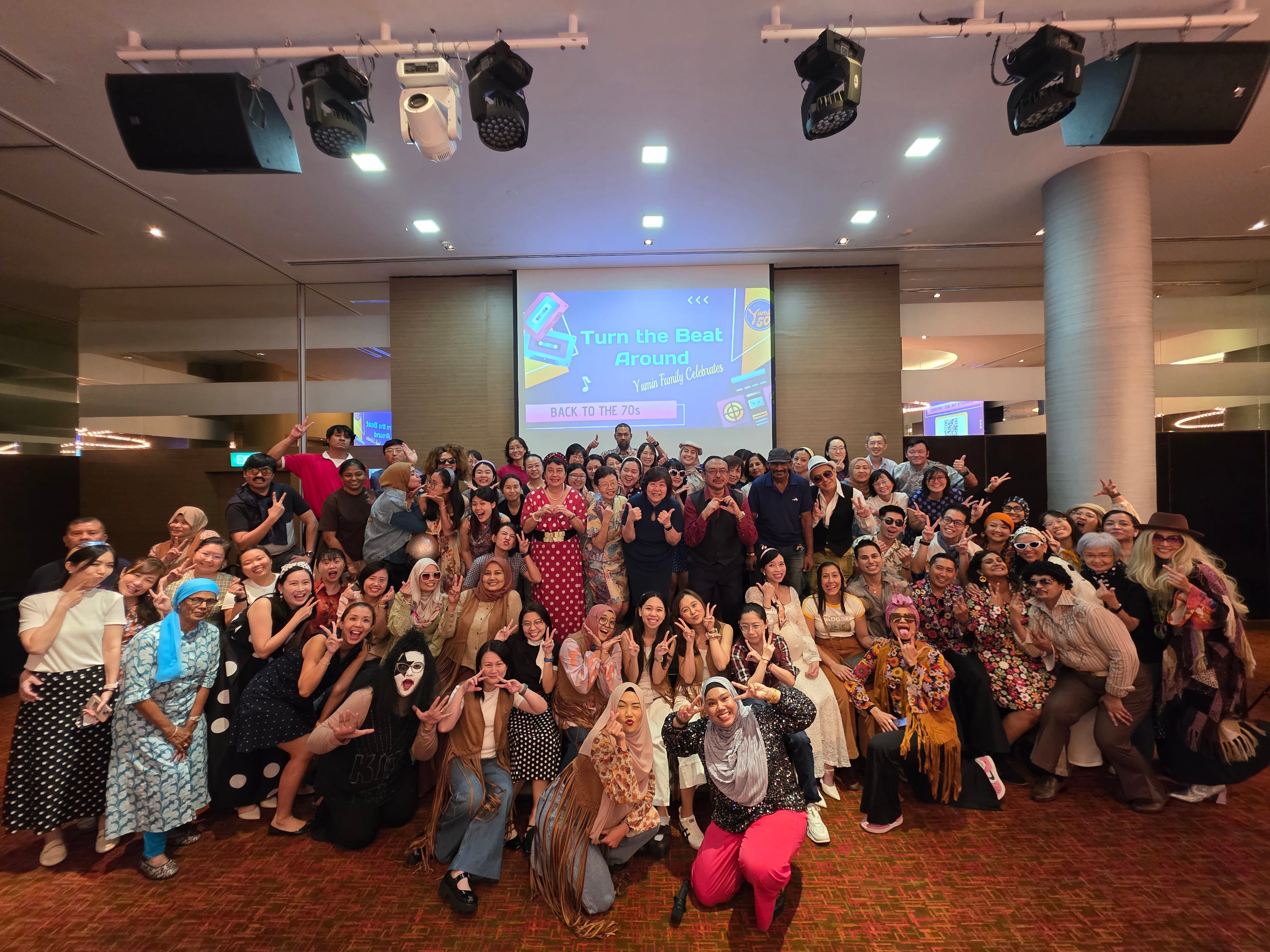 Large group in 70s costumes posing at a "Turn the Beat Around, Back to the 70s" themed party indoors.