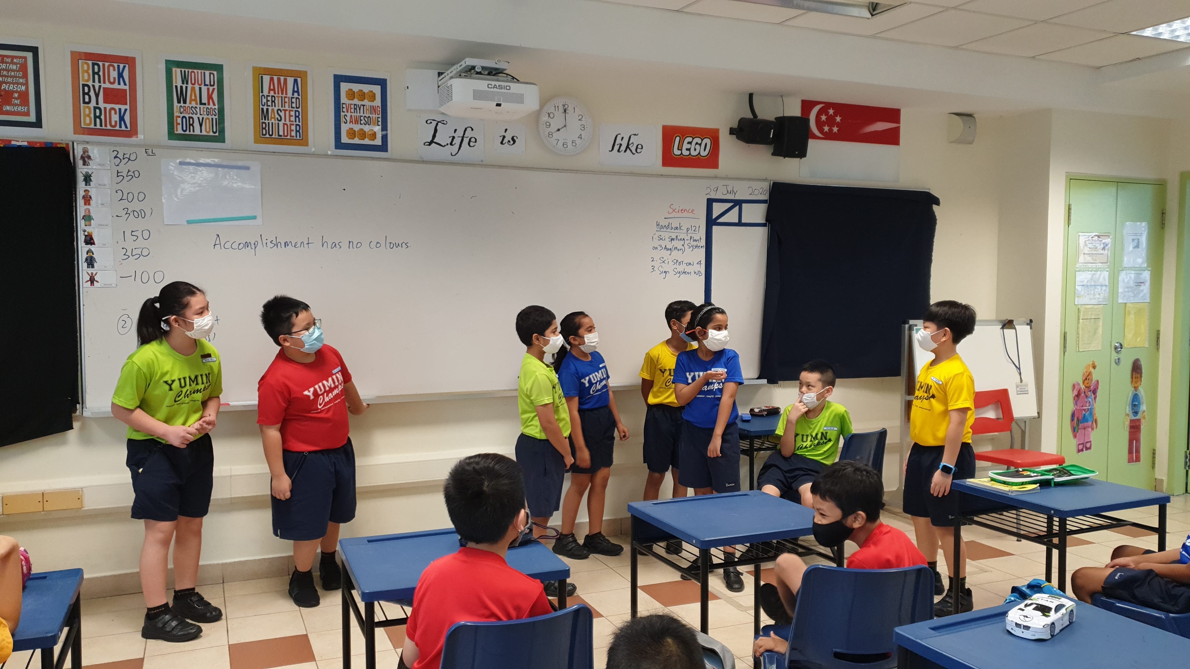 Children wearing masks and colorful shirts stand and sit in a Lego-themed classroom with a whiteboard behind them.