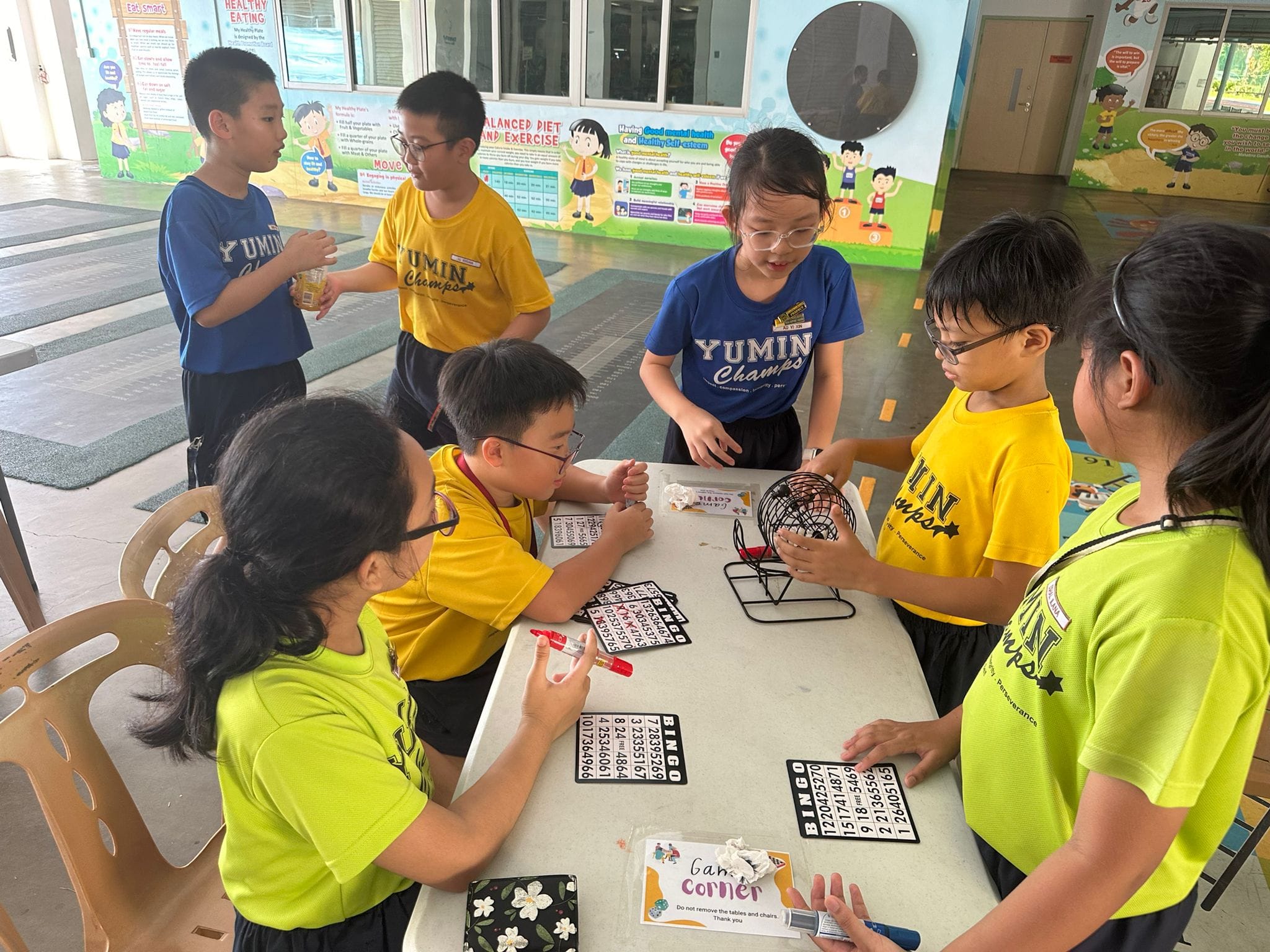 Students play bingo at a table with a wire cage tumbler and bingo cards.