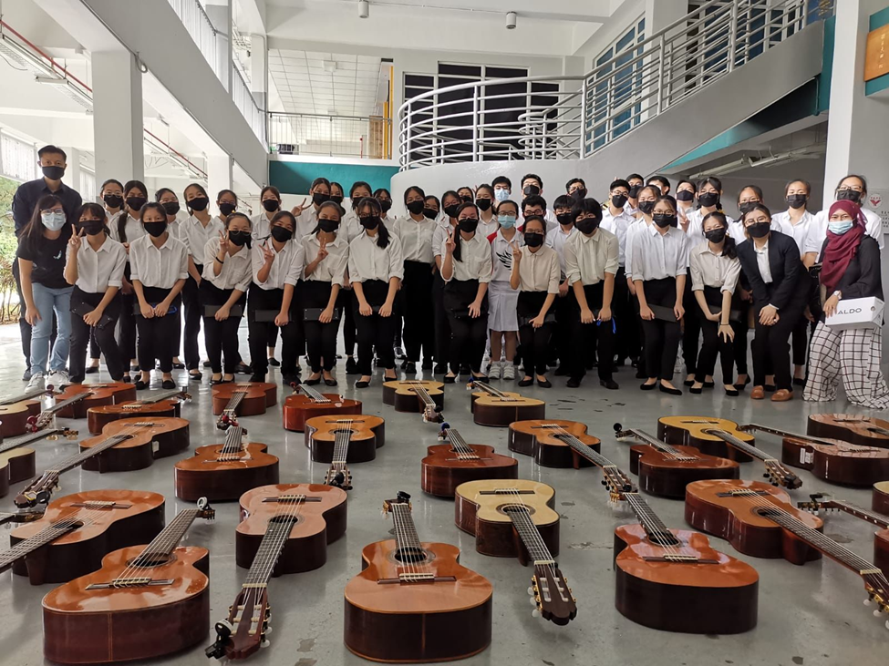 Large group of masked students in white shirts posing indoors behind many acoustic guitars laid on the floor.