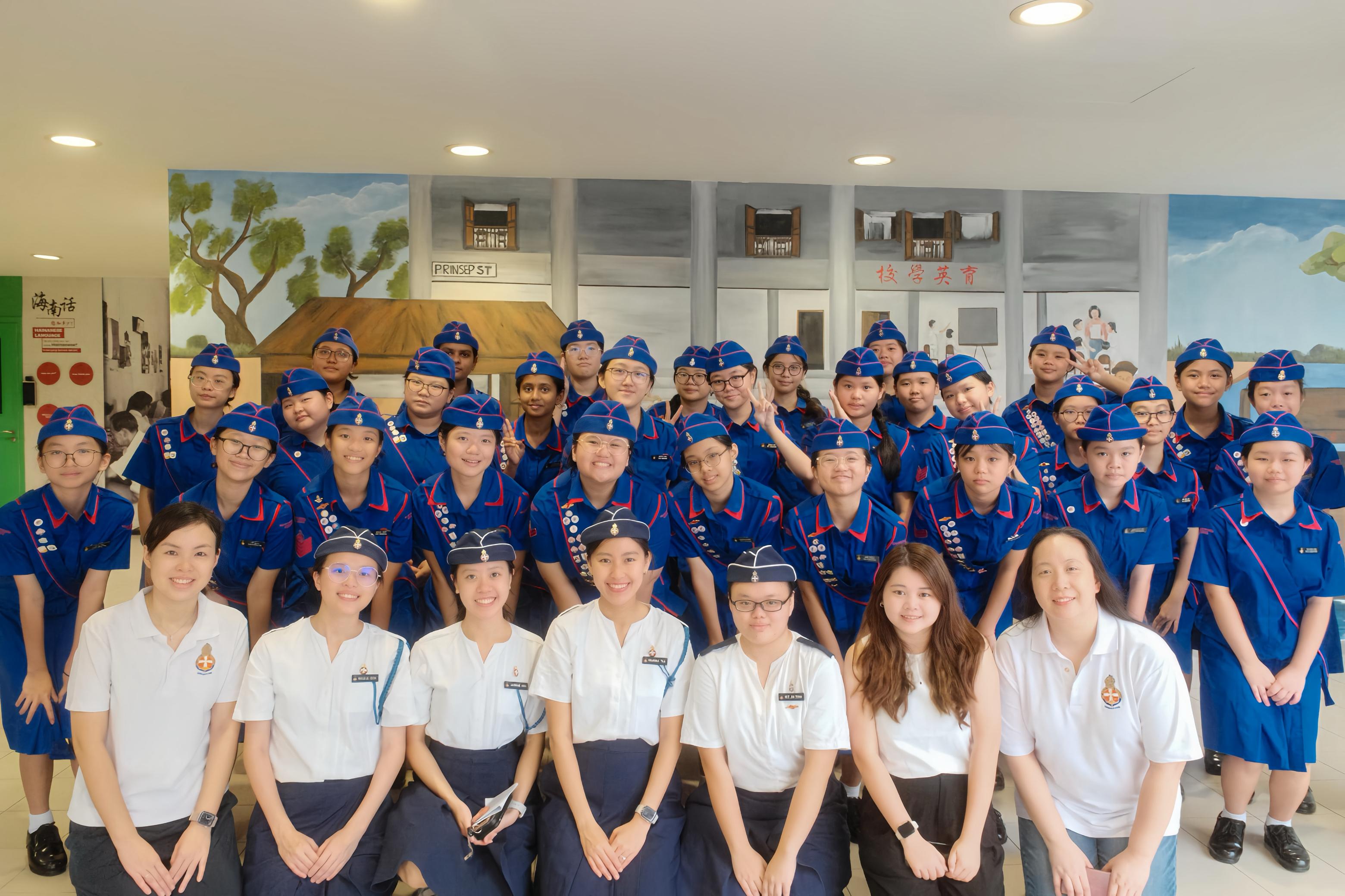 Large group of young students in blue uniforms with red trim and caps posing indoors in front of a painted mural.