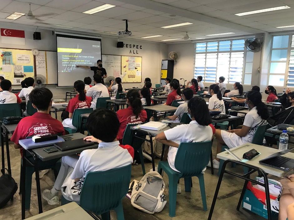 Students in white and red uniforms sit at desks facing a teacher presenting slides on a projected screen.