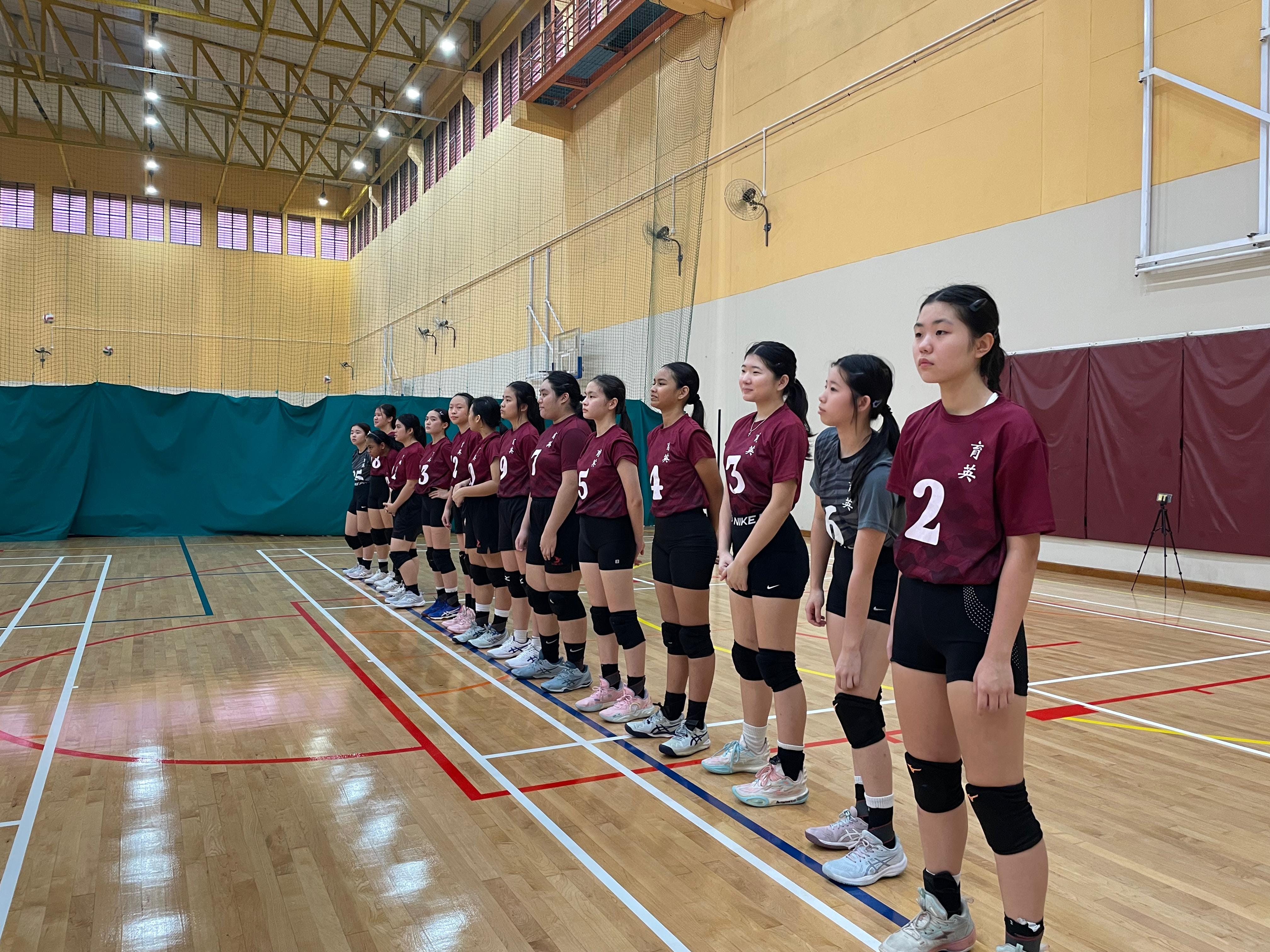 A row of young female volleyball players in dark red numbered jerseys standing in line on an indoor wooden court.