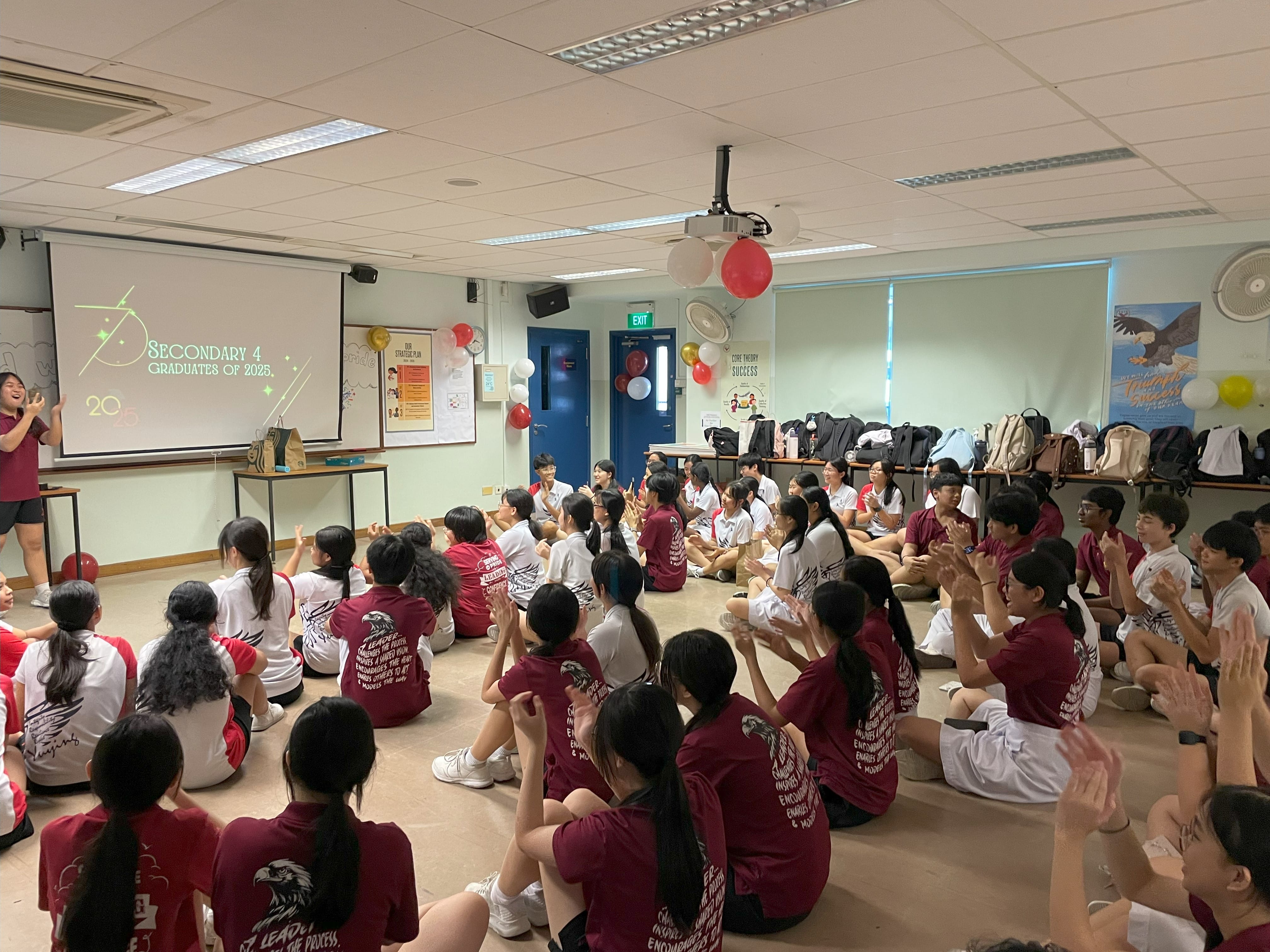 Students in maroon and white uniforms sit on a floor clapping during a Secondary 4 Graduates of 2025 celebration.