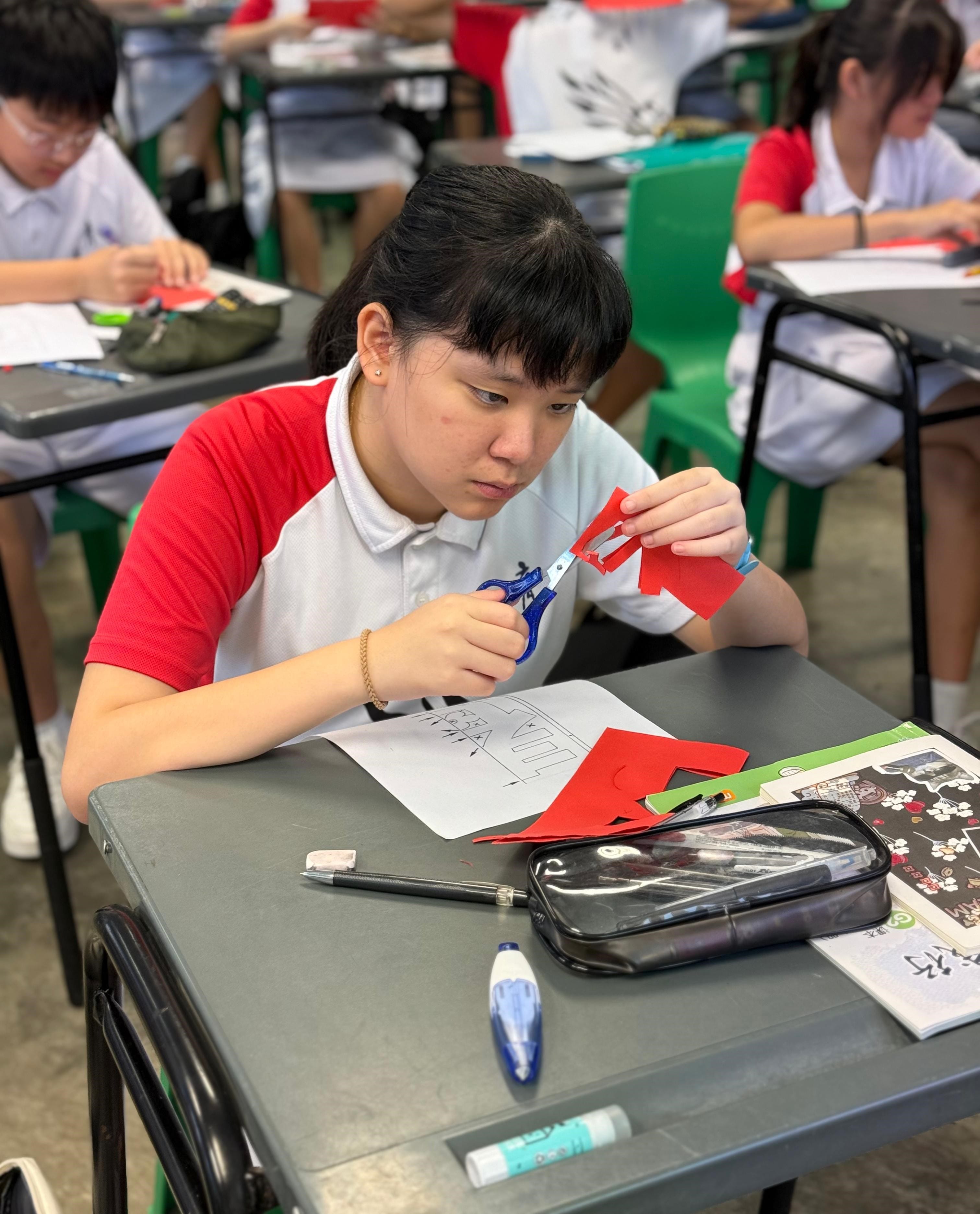 A student in a red and white uniform cuts red paper with scissors at a desk in a classroom.