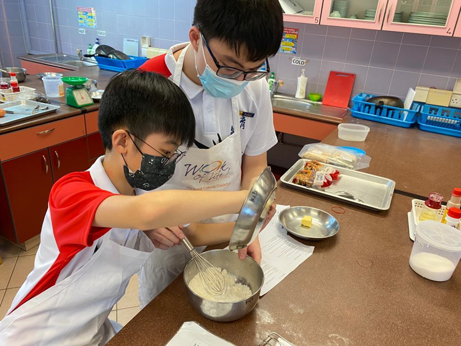 Two masked boys wearing aprons whisk batter in a steel bowl during a cooking class.