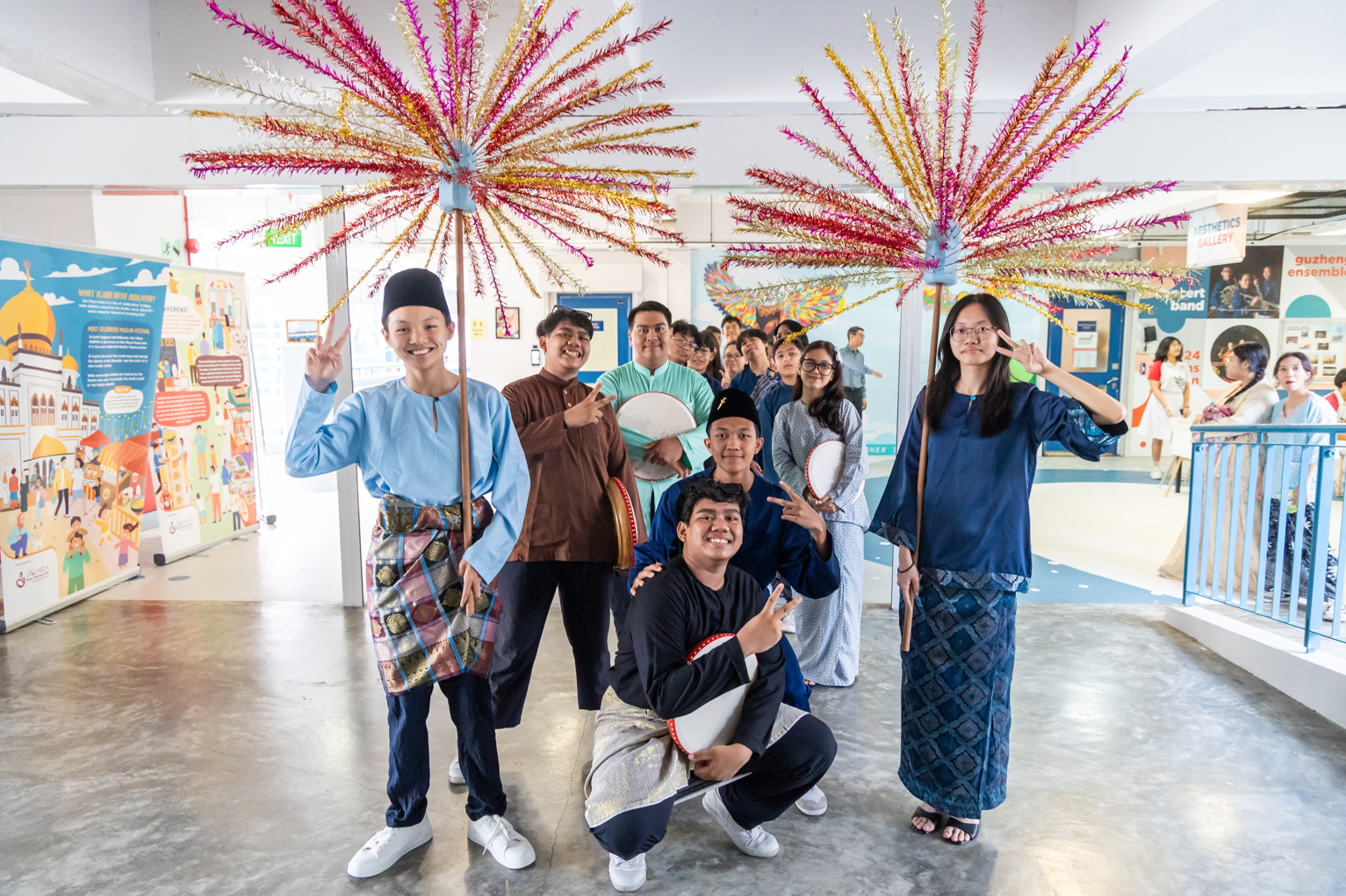 Students in traditional Malay attire pose with colorful decorative umbrellas and drums indoors at a cultural event.
