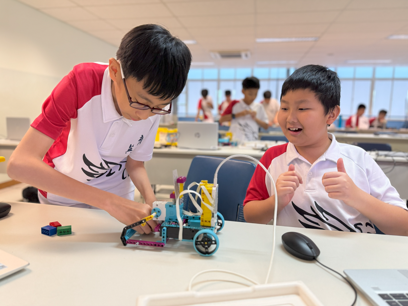 Two boys work on a colorful wheeled robot at a desk; one gives a thumbs up while the other adjusts the build.