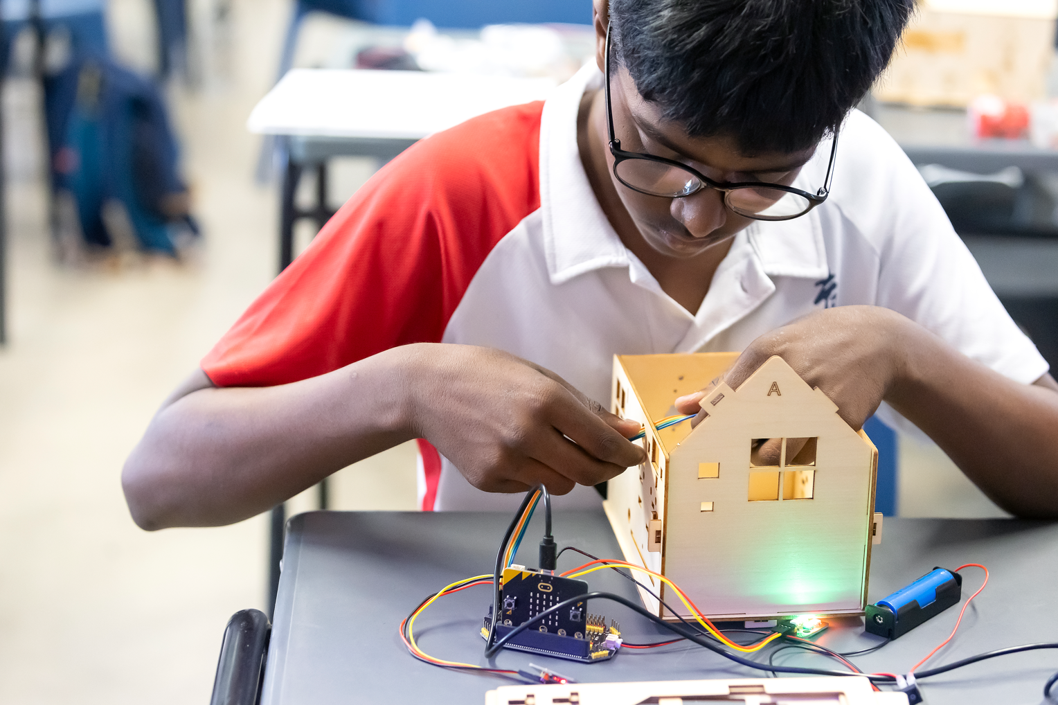 A boy wearing glasses wires a small wooden house model connected to a microcontroller with colorful cables on a desk.