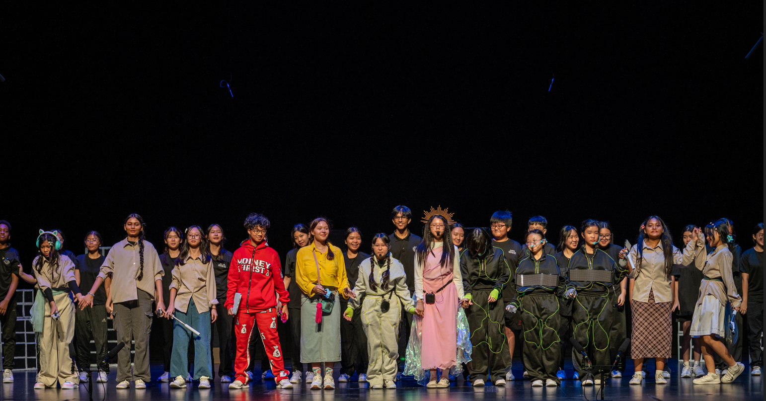 A large group of young performers in varied costumes stand together on a dark stage after a show.