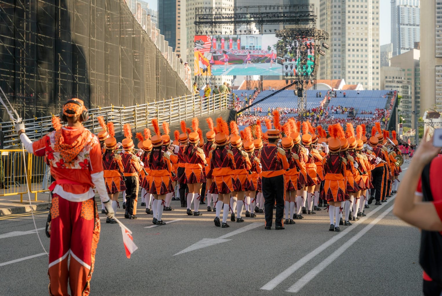 A marching band in bright orange uniforms with feathered hats parades down a street toward a large outdoor event venue.