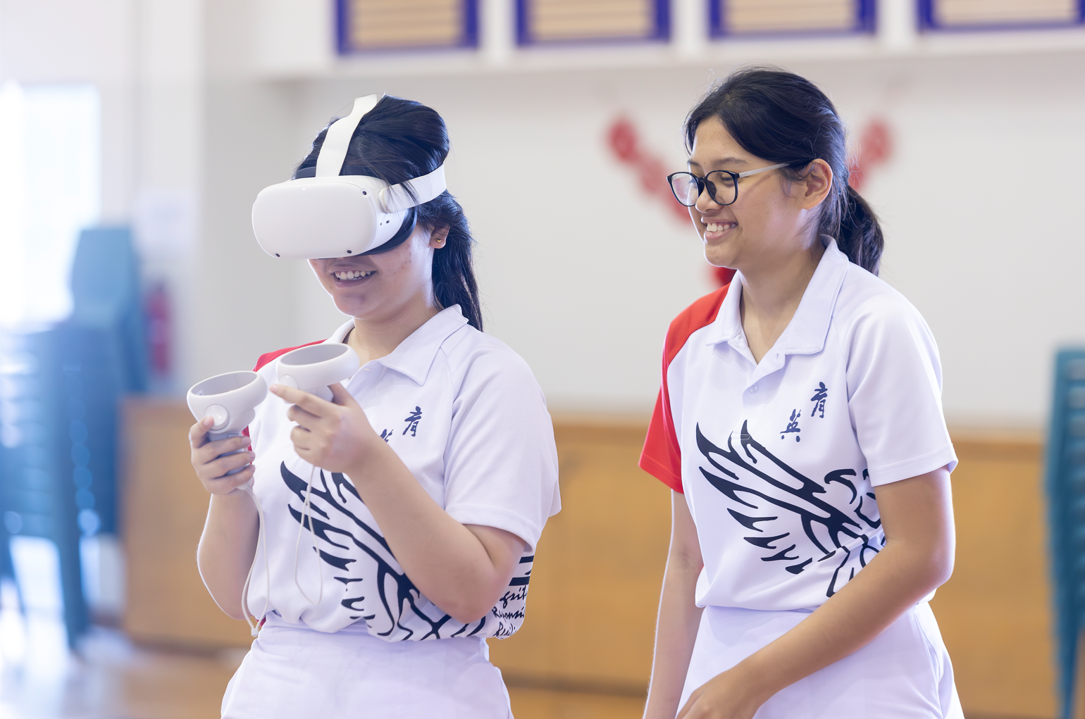 Two young girls in white uniforms, one wearing a VR headset and holding controllers, smiling in a school gym.