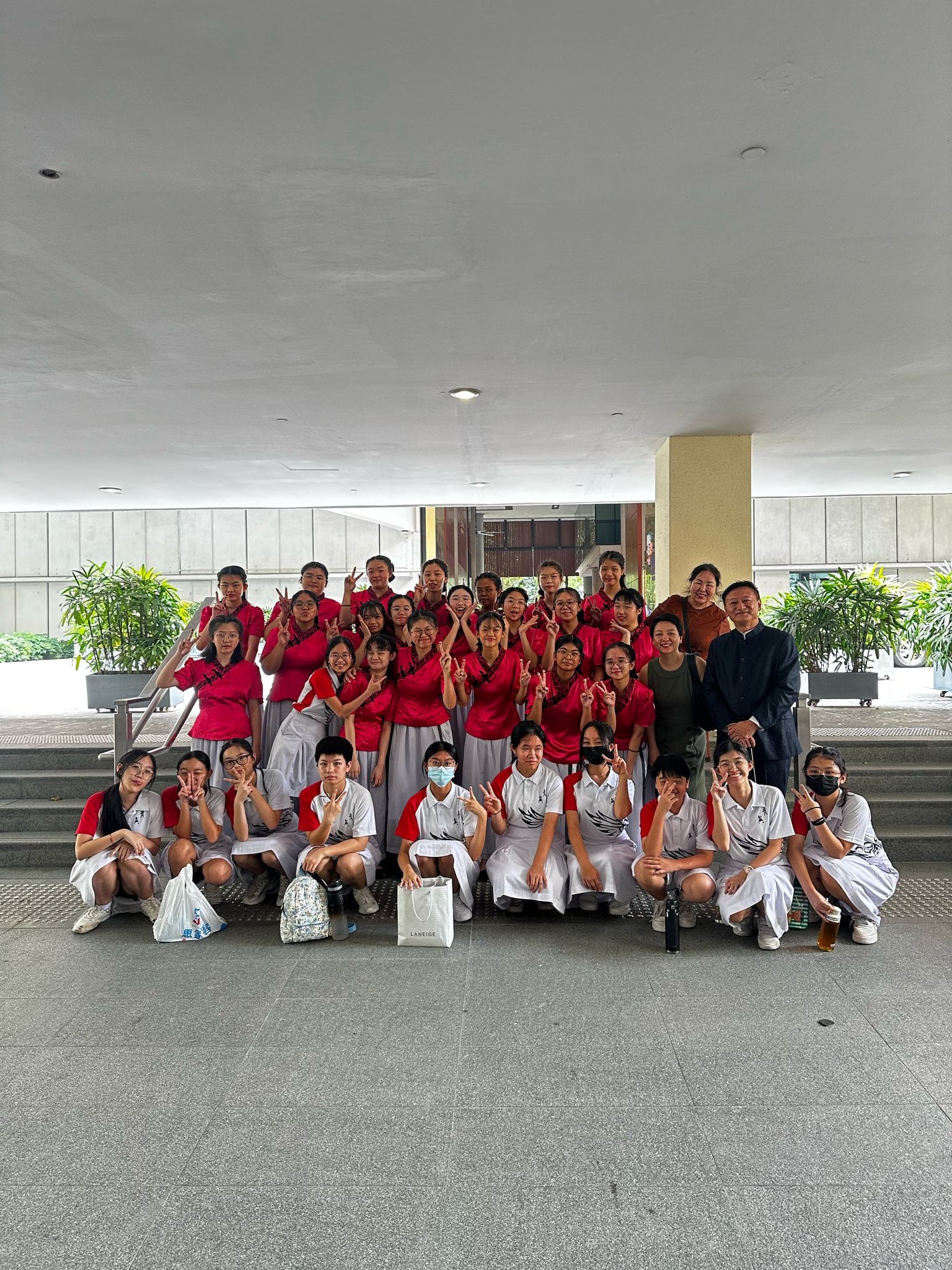 A large group of students in red and white uniforms pose with peace signs on steps under a covered area.