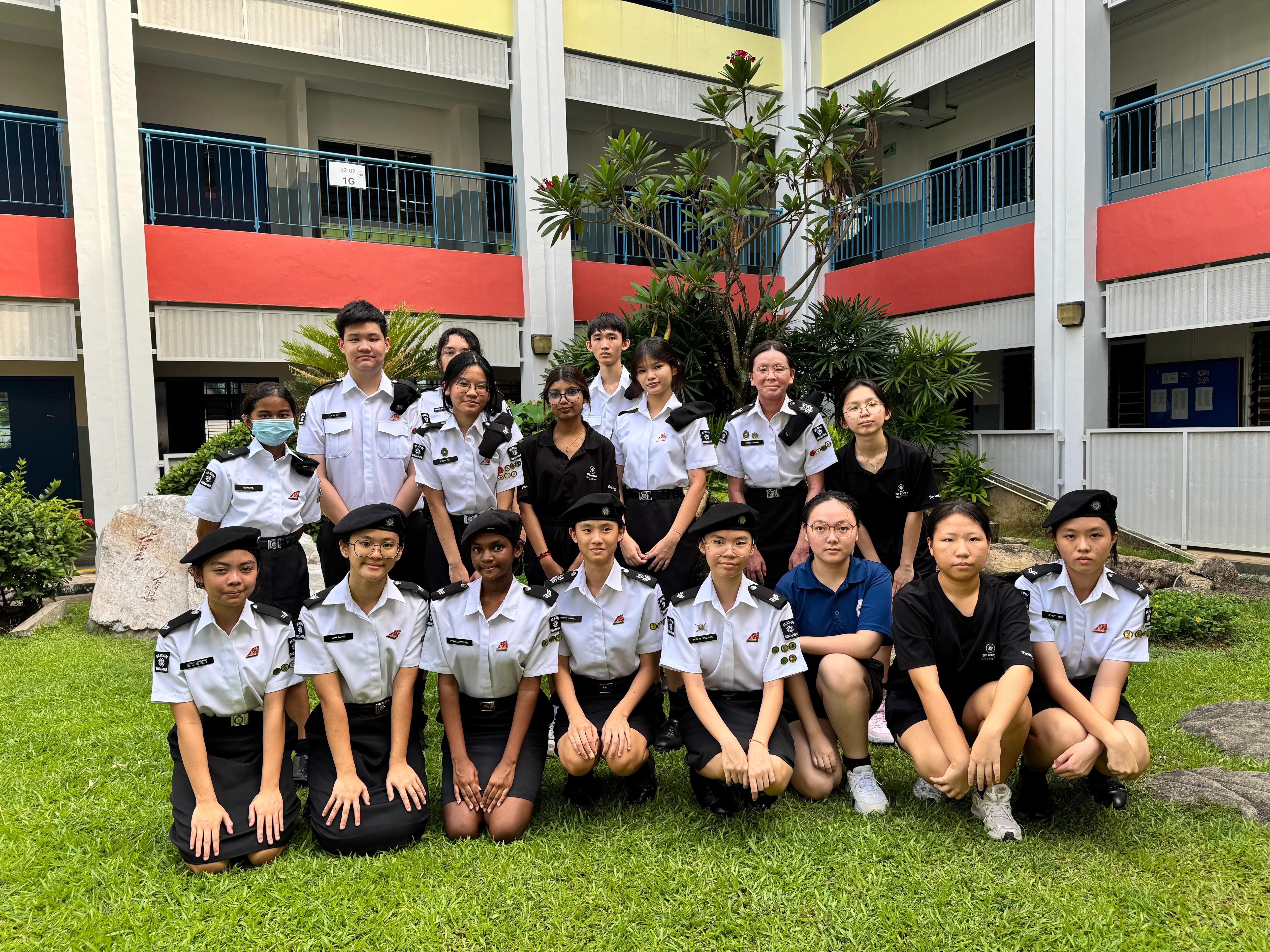 A group of students in white and black uniforms with berets pose on grass in a school courtyard.